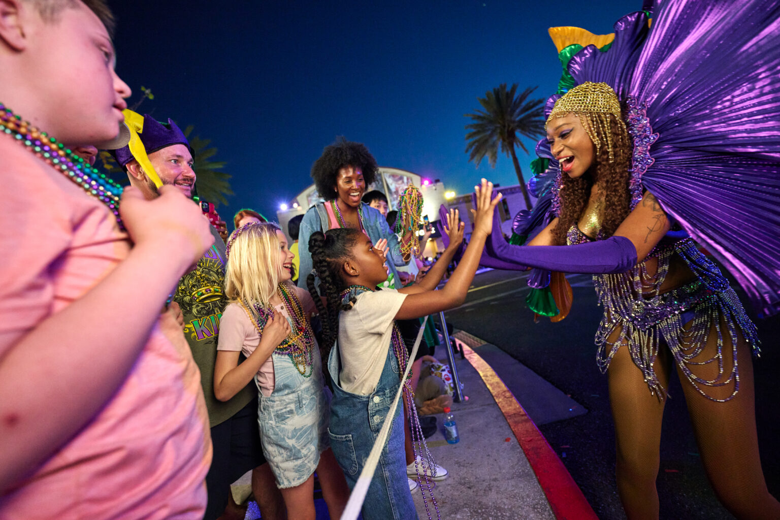 A night parade scene at dusk, with a performer in a huge fanned purple costume and a gold beaded headpiece reaching in from the roadway to high five a young girl in denim overalls and bead strands, while two other children and a man in a purple jester hat watch, palm trees and a lit float behind them against a deep blue sky
