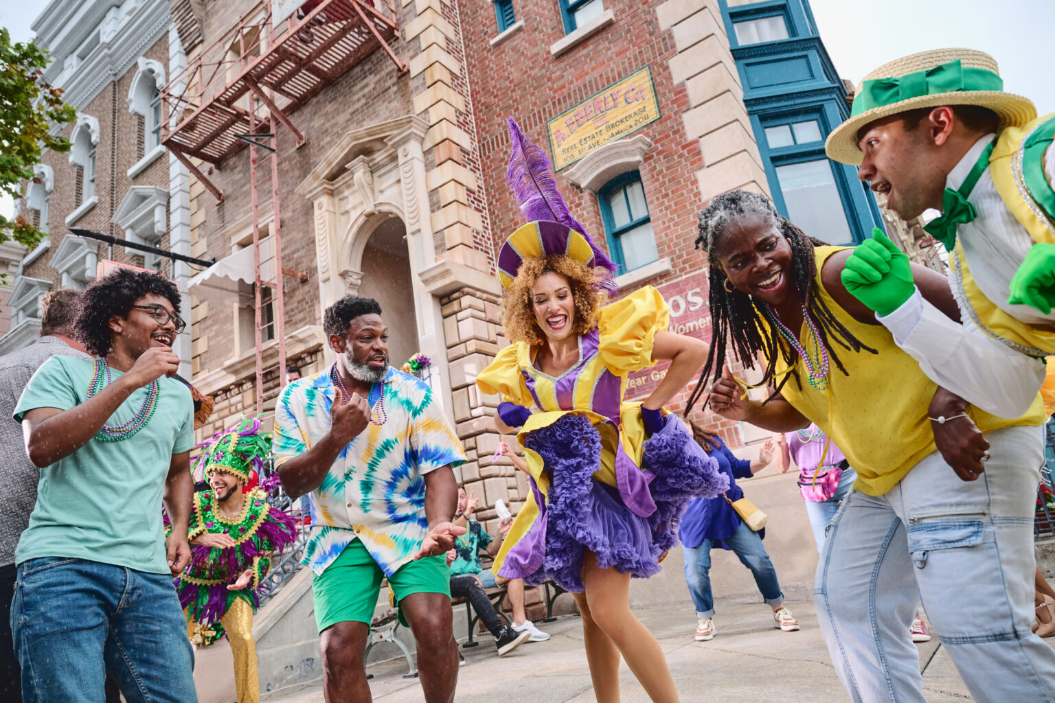 A daytime street party on the New York backlot, with a performer in a purple and gold ruffled dress and a tall purple feathered headdress dancing at the center, surrounded by laughing guests in strands of green, gold and purple beads, and a man in a straw boater hat with a green bow tie and green gloves at the right, in front of a brick building with a fire escape and an EBERLY Co. Real Estate Brokerage sign