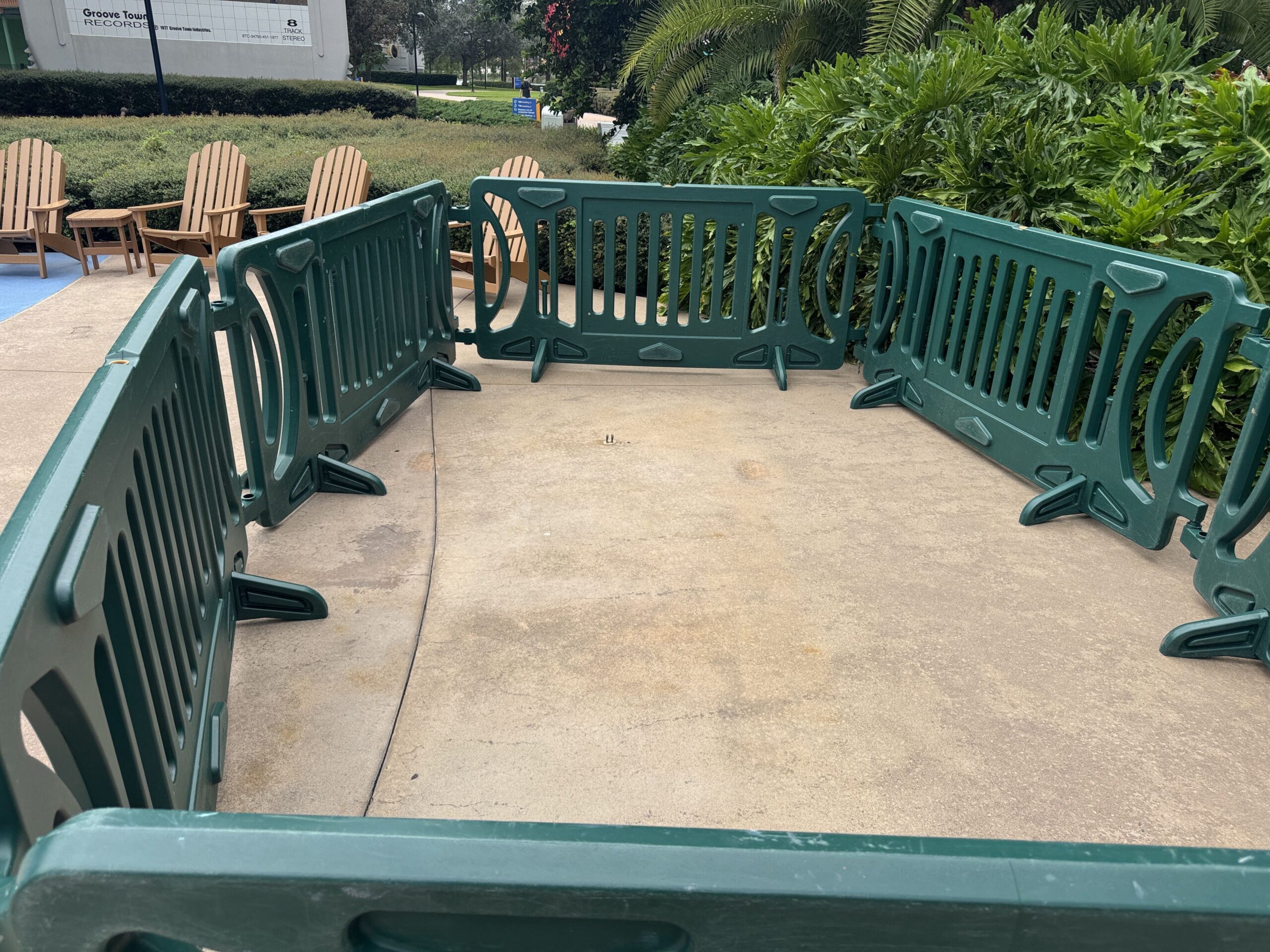 An empty rectangle of concrete fenced in by green plastic barricades, with Adirondack chairs behind it and the giant Groove Town Records 8 track tape in the background
