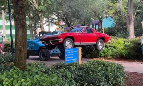 A red classic style hot rod lifted off the ground on the front of a blue utility vehicle, with Pop Century landscaping, a blue wayfinding sign and trees around it