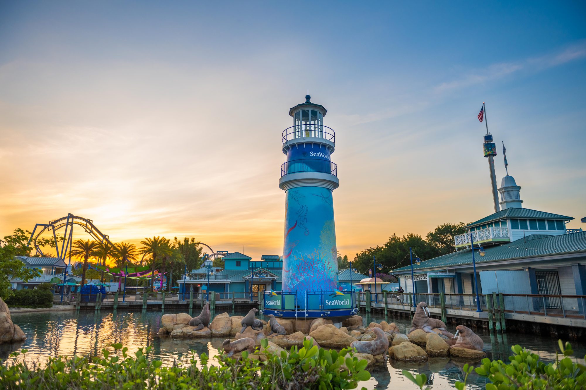 SeaWorld Orlando's blue entrance lighthouse at sunset, painted with a seahorse, fish and coral and topped with a lamp room and railing, standing on rocks in the entrance lagoon surrounded by sculpted sea lions and walruses, with palm trees and a roller coaster silhouetted against an orange sky at the left and the Sky Tower and a VIP Entrance building at the right