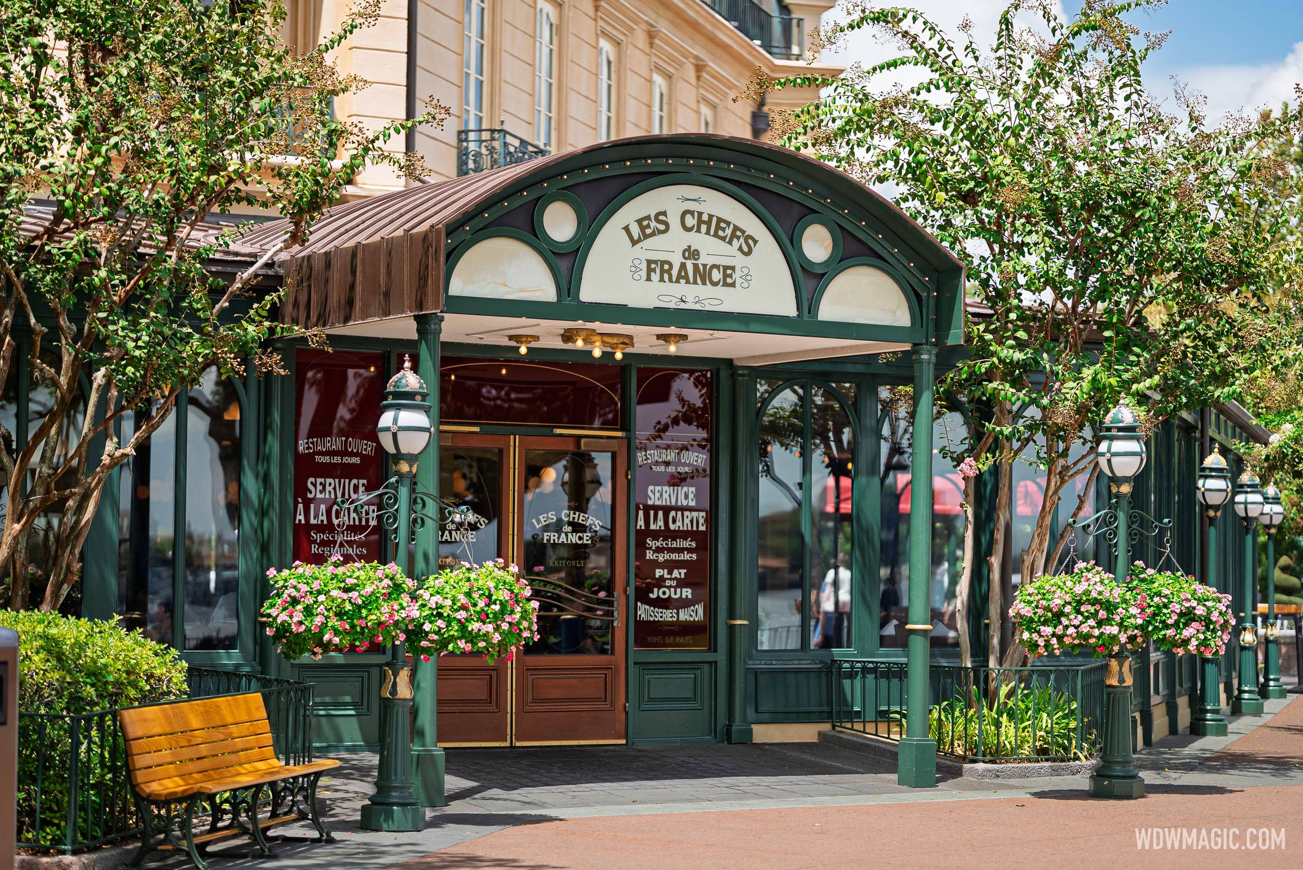 An arched brown metal canopy over an exit-only door of Les Chefs de France with its green frame, cream sign, hanging baskets of pink flowers, lamp posts and dark red window posters reading Service a la Carte