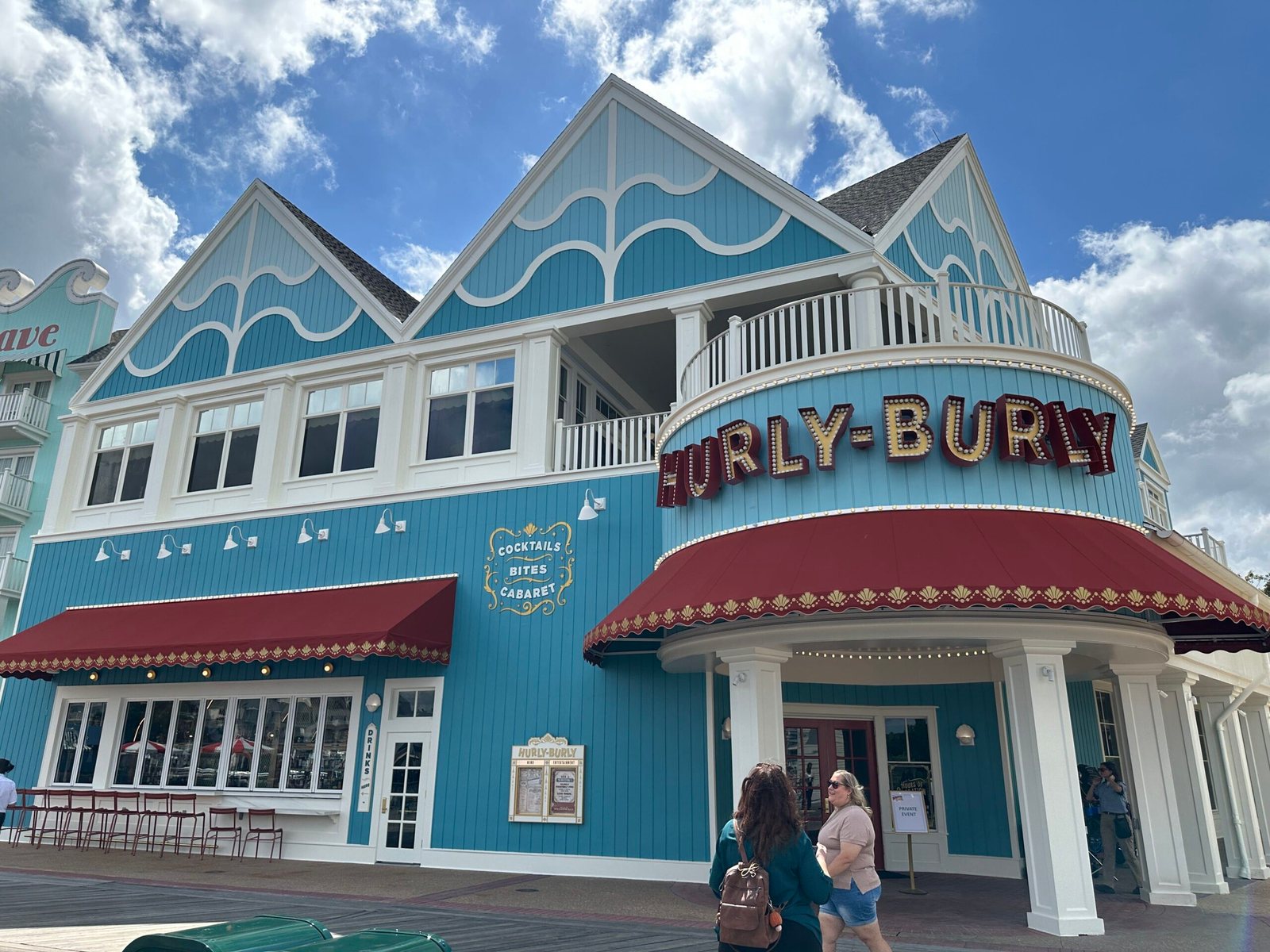 The Hurly-Burly building on Disney's BoardWalk: a teal board-and-batten facade with a curved red awning, a HURLY-BURLY marquee lined with bulbs and a painted Cocktails, Bites, Cabaret sign, with guests walking past on the boardwalk