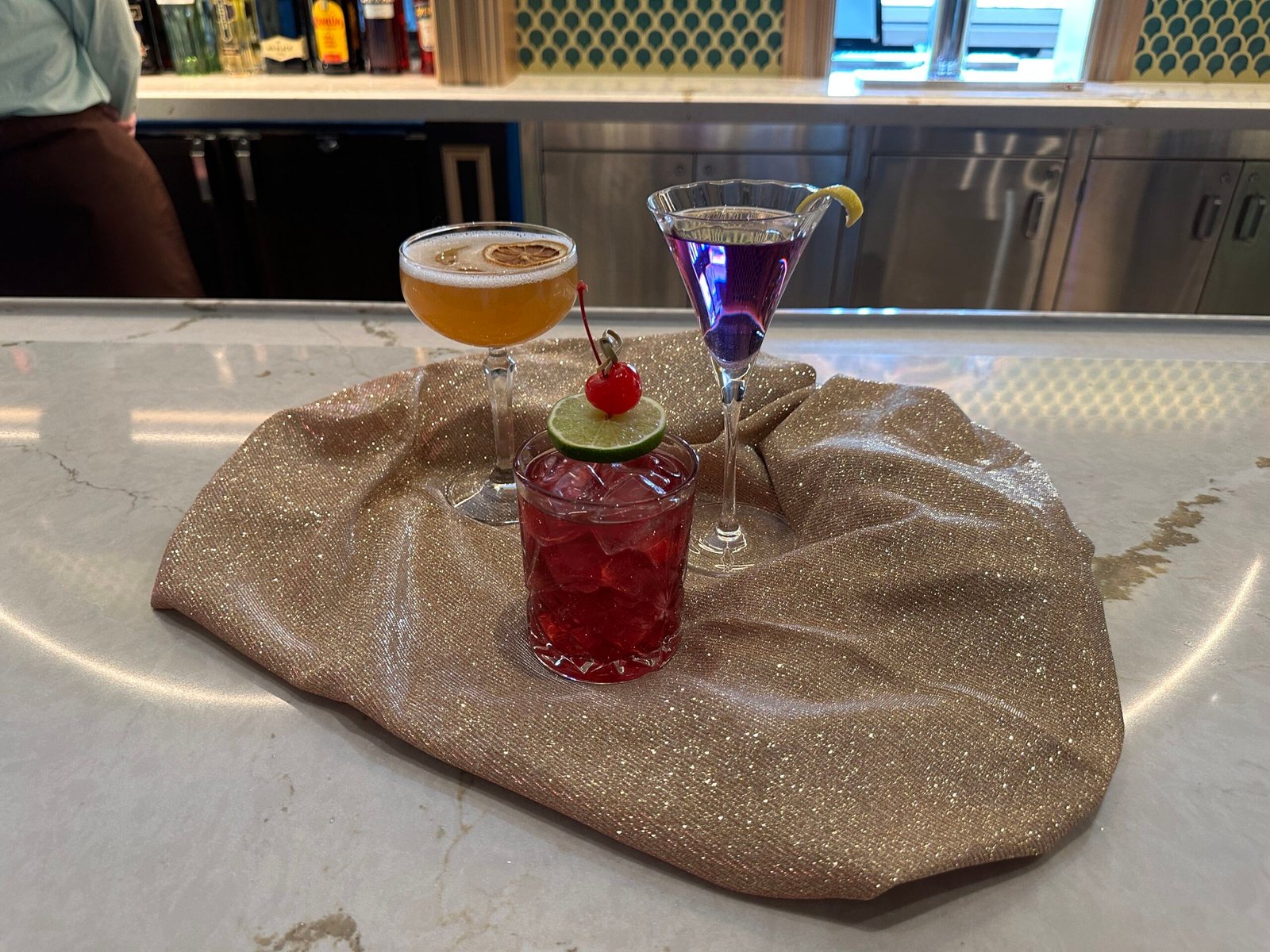 Three cocktails on a sparkly gold cloth on a marble bar: a foamy whiskey sour in a coupe, a red drink with a cherry and lime wheel in a rocks glass, and a purple martini with a lemon twist