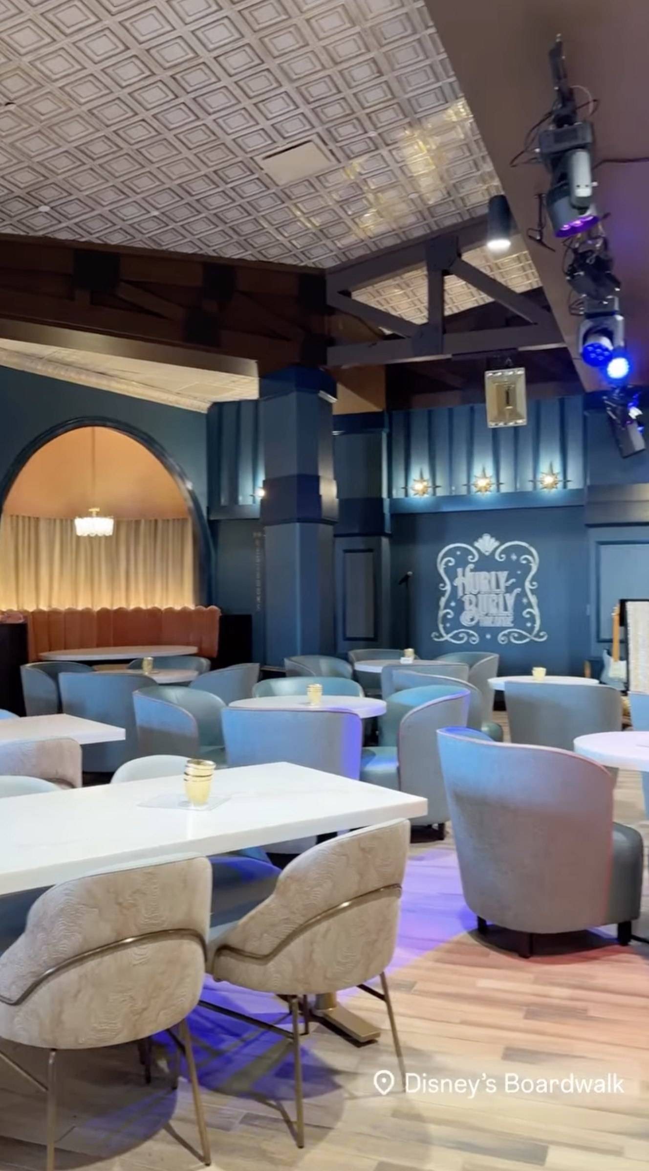 The Hurly-Burly Theatre logo in white on a blue wall, beside rows of grey and blue tub chairs, white tables and a pressed tin ceiling, with an arched booth alcove and chandelier at left