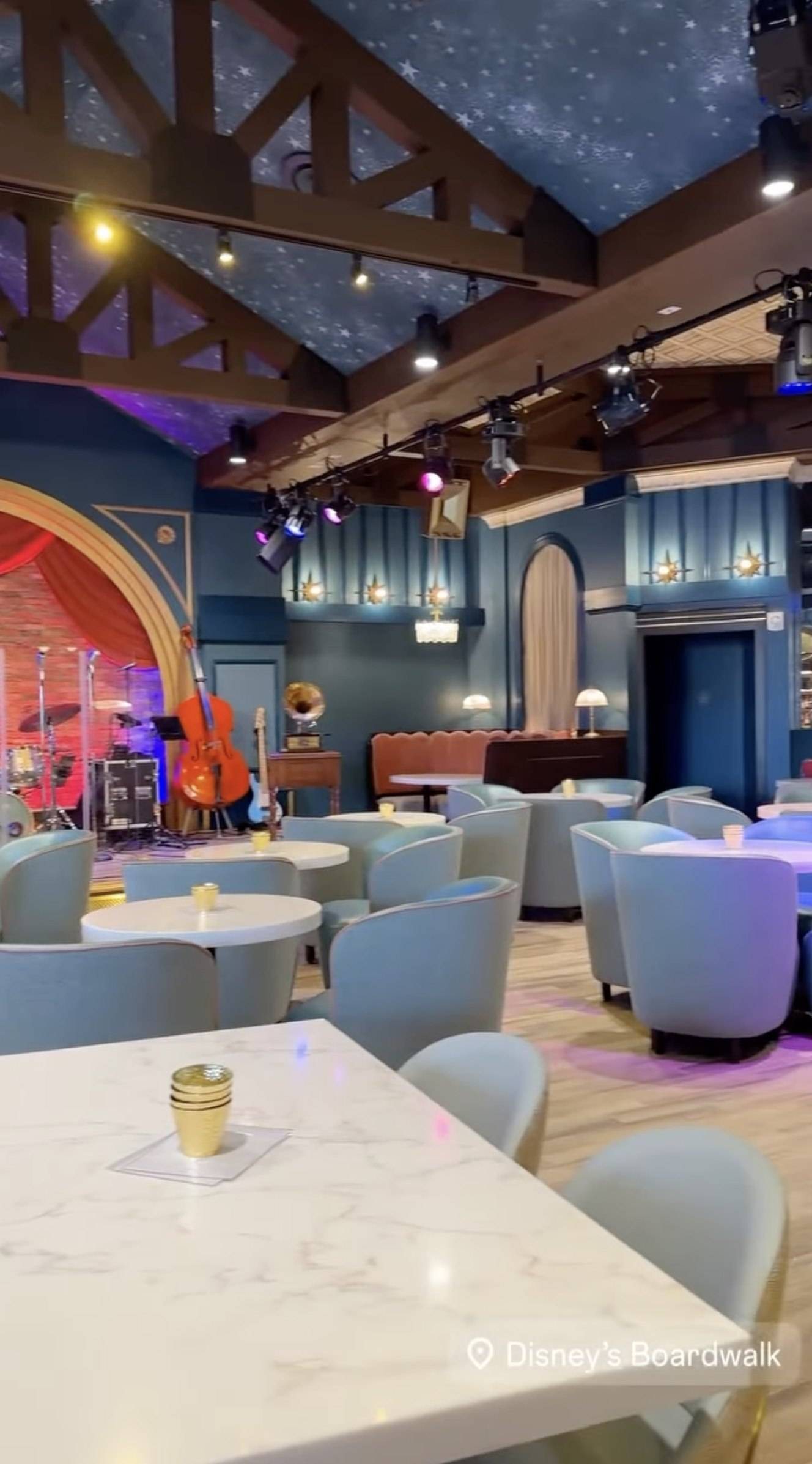 Another angle inside Hurly-Burly: blue tub chairs and white tables in the foreground, the stage with an upright bass and a gramophone at left, and a curved booth under an arched alcove in the back