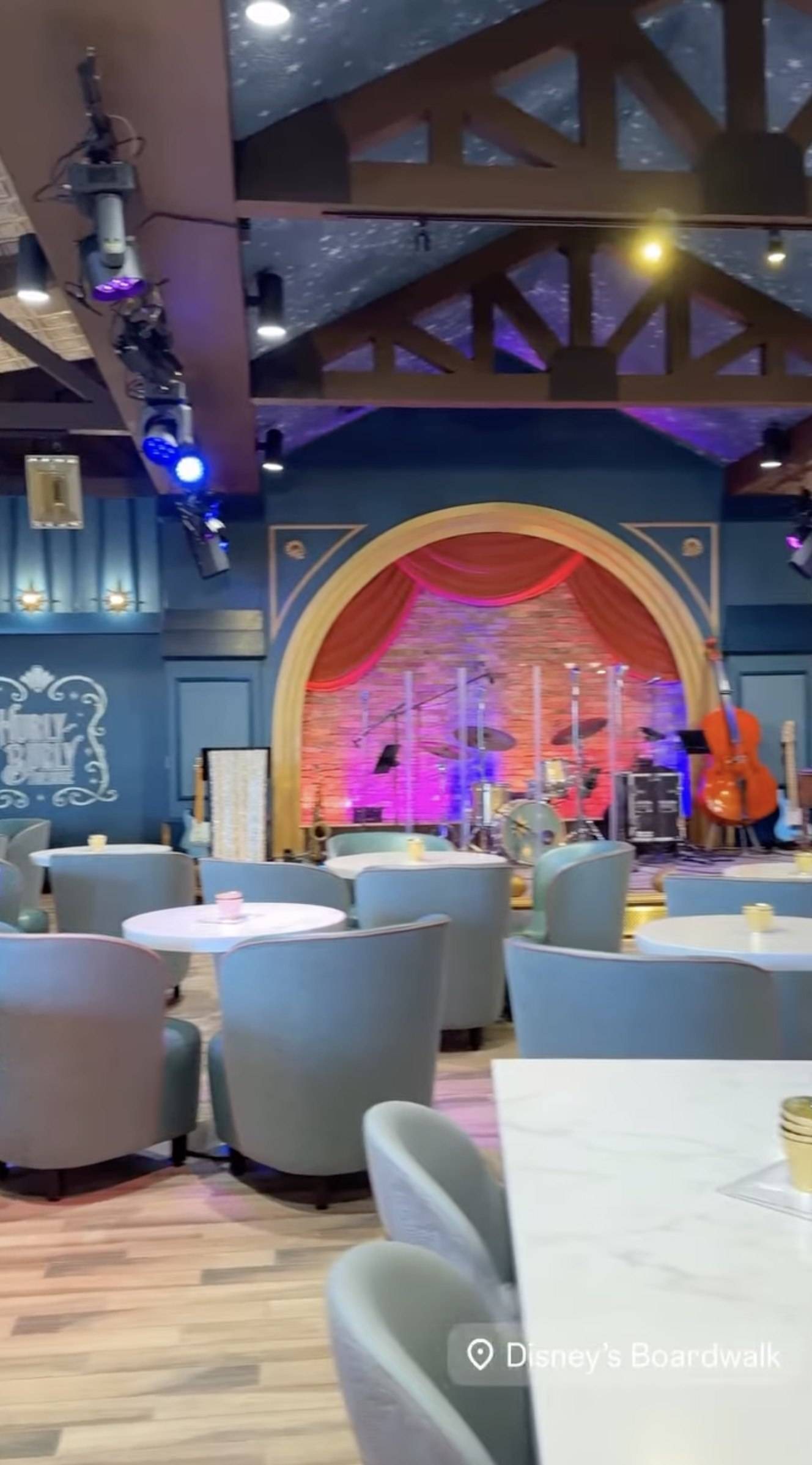 Inside Hurly-Burly: rows of powder blue tub chairs around white tables facing a gold-arched stage with red curtains, a brick back wall, drums, an upright bass and guitars, under wooden ceiling trusses and a starry ceiling