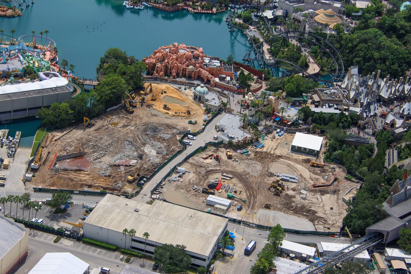 Aerial view of The Lost Continent construction site: two large cleared dirt areas with equipment, the Islands of Adventure lagoon at the top, and the orange rockwork of Mythos with the blue-green dome of Treasures of Poseidon just below it