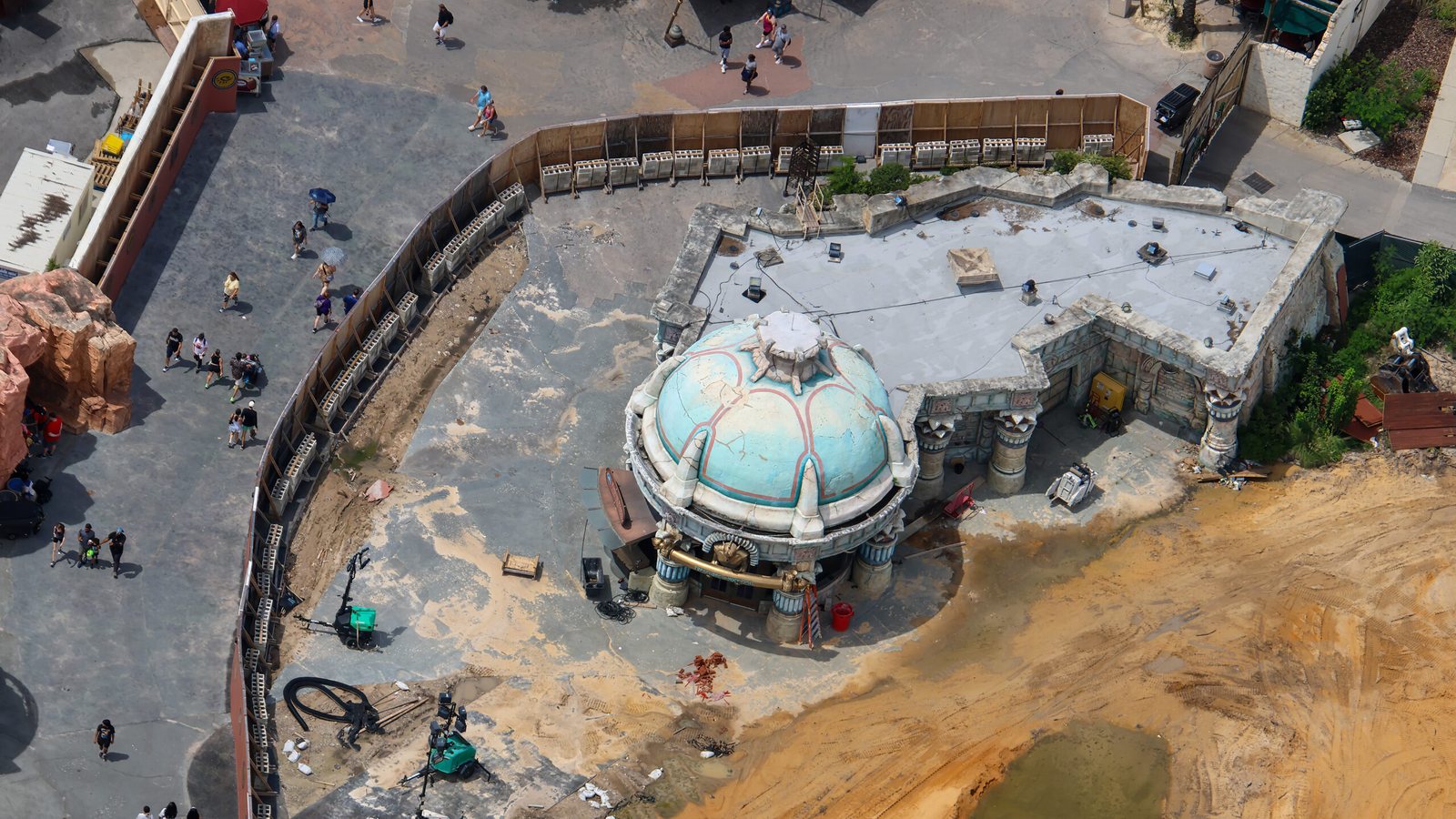 Aerial view of Treasures of Poseidon's blue-green dome and flat roof still standing, surrounded by construction walls and a cleared dirt site, with guests on the walkway at left