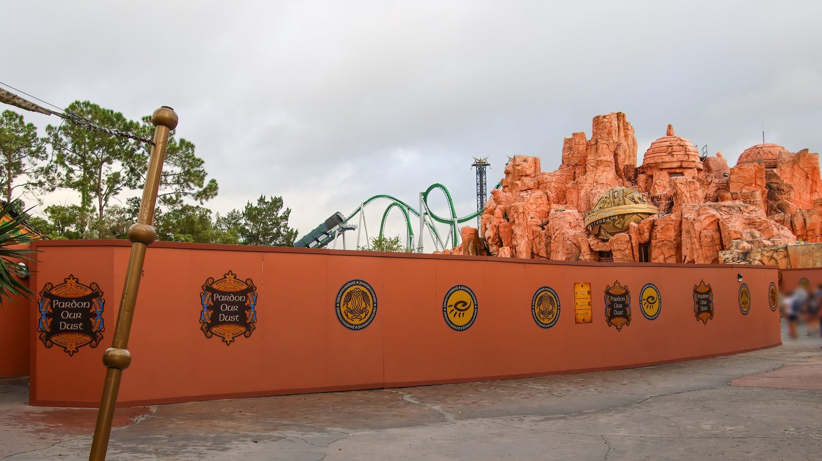 An orange construction wall with Pardon Our Dust medallions curving along a walkway in The Lost Continent, with the green track of The Incredible Hulk Coaster visible over the wall and the orange rockwork of Mythos Restaurant on the right
