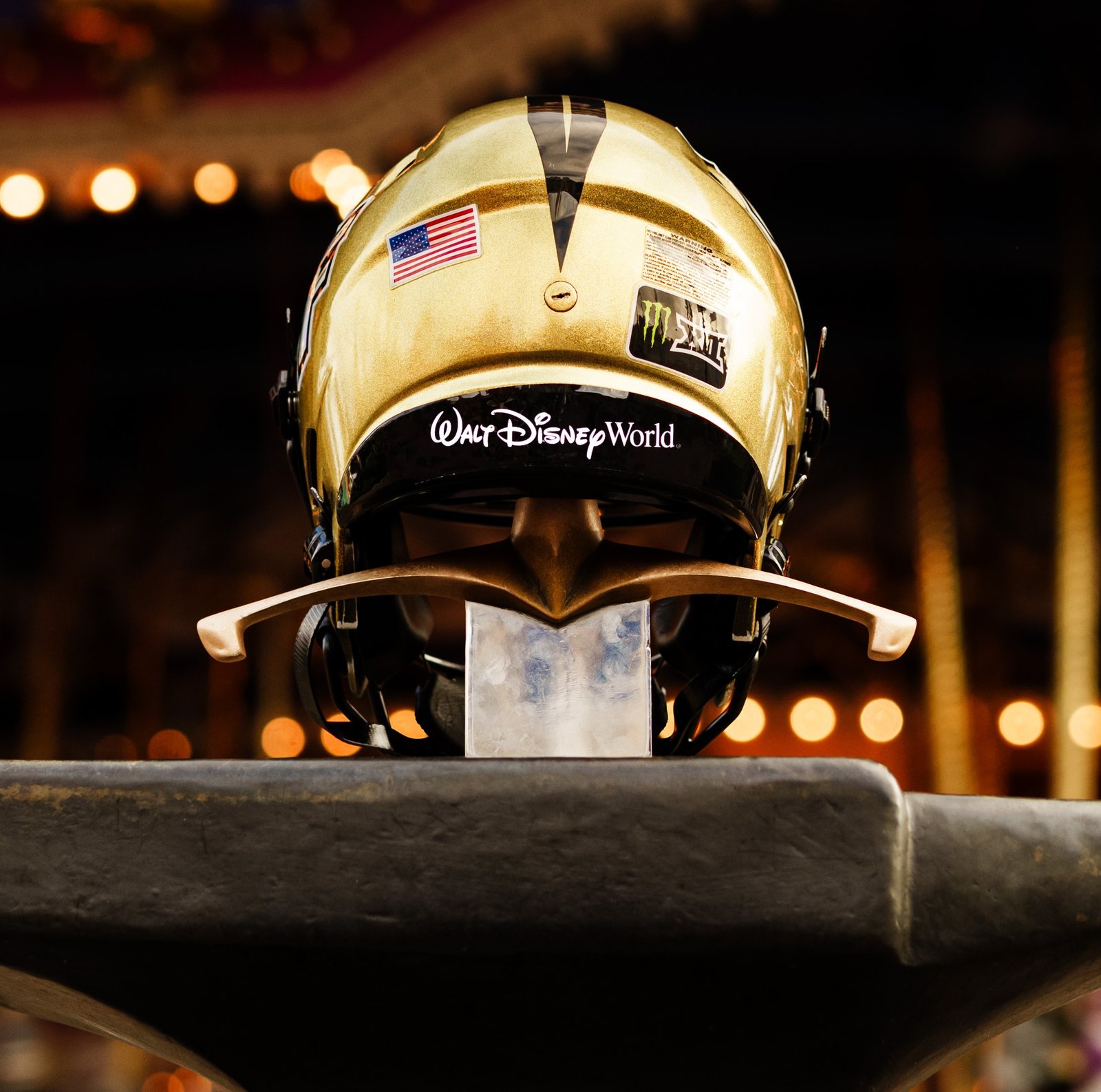 The back of the gold UCF helmet, with the Walt Disney World script on its bumper, resting on the hilt of a sword set in a dark anvil, with blurred warm lights in the background