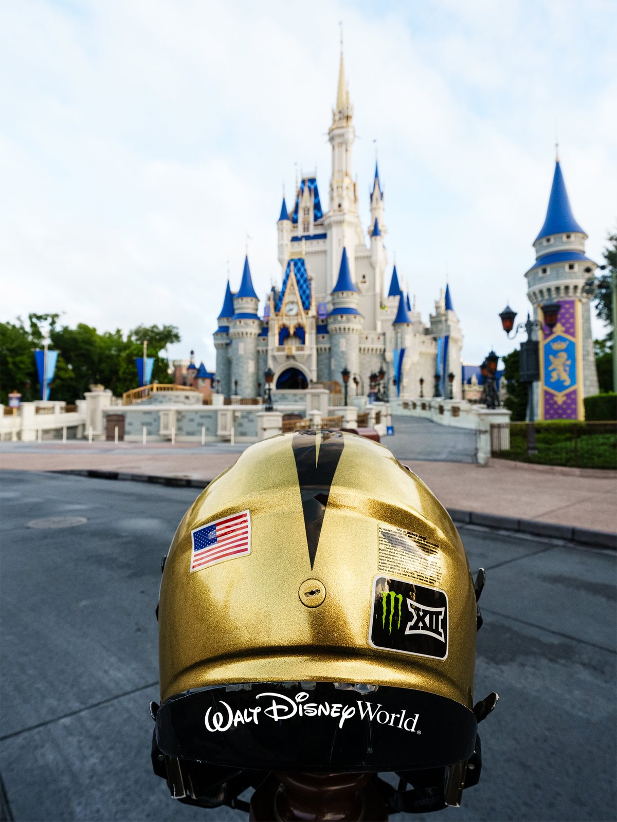 The back of a gold UCF football helmet with an American flag decal, a Monster Energy and Big 12 sticker and the white Walt Disney World script on its black rear bumper, with Cinderella Castle in the background