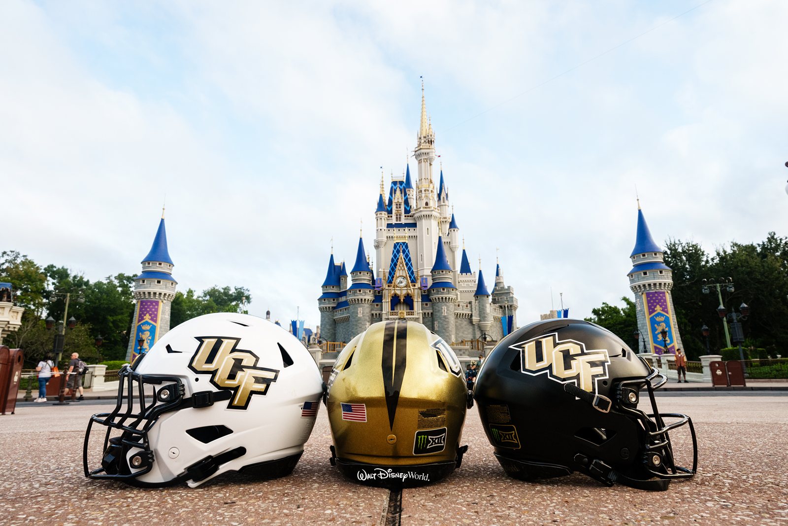 Three UCF football helmets, white, gold and black, lined up on the pavement in front of Cinderella Castle, with the Walt Disney World script visible on the rear bumper of the gold helmet in the middle