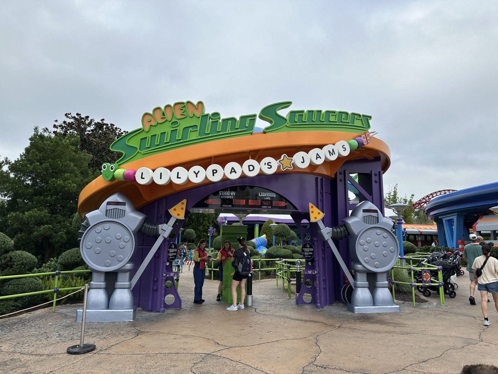 The Alien Swirling Saucers entrance arch with a row of round white letter beads spelling LILYPAD'S JAMS under the ride's green and orange sign, flanked by two gray robot statues, with guests walking in