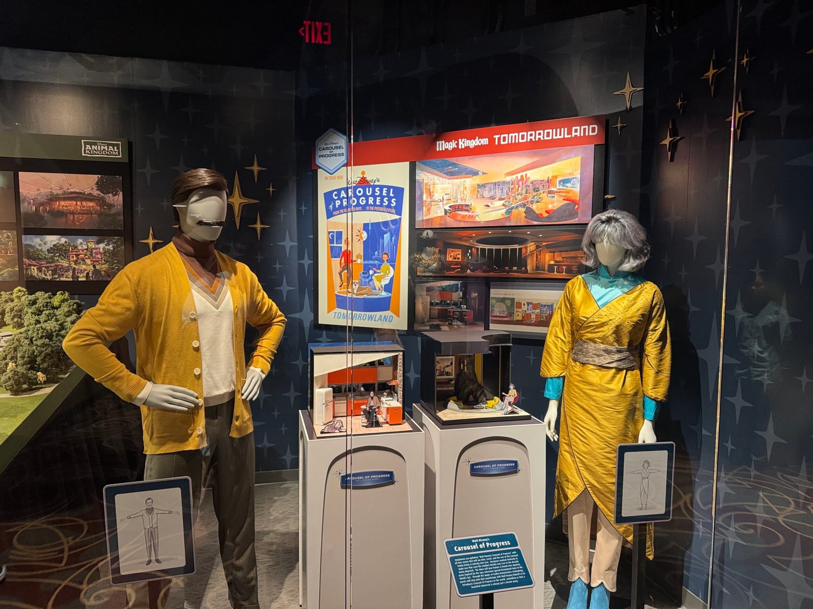 Two mannequins against a starry blue wall: at left, a man's costume with a mustard yellow cardigan over a white V-neck and brown turtleneck, with dark trousers; at right, a woman's yellow dress with a blue collar. Between them, a Carousel of Progress poster, a Tomorrowland sign and small scale dioramas on white pedestals