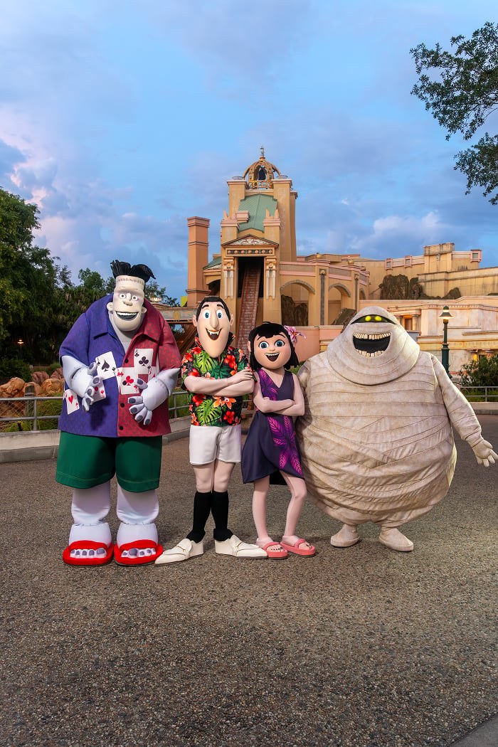 Costumed Hotel Transylvania characters Frank, Drac, Mavis and Murray the mummy posing together in summer outfits on a SeaWorld Orlando walkway, with a tan tower and trees behind them