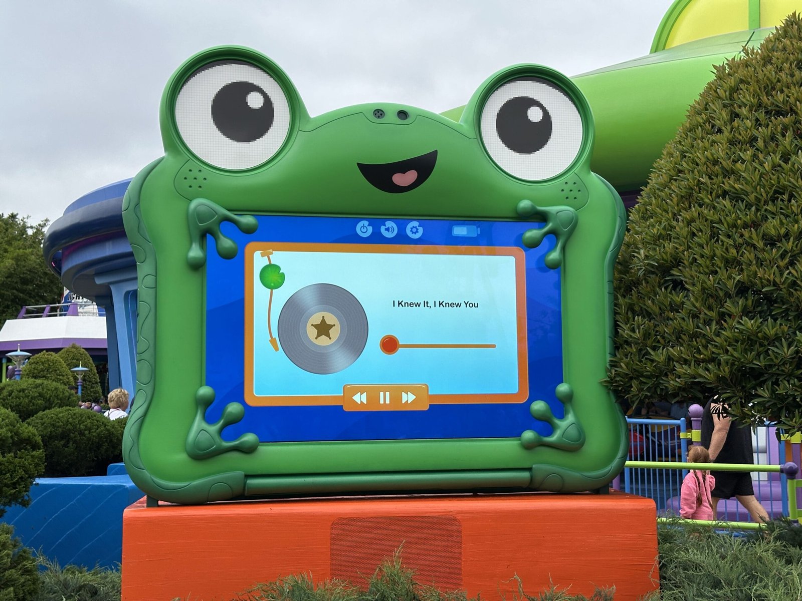 Lilypad, a large green frog-shaped tablet with big cartoon eyes and a smile, standing on an orange base outside Alien Swirling Saucers, its screen showing a record player app with the track title I Knew It, I Knew You