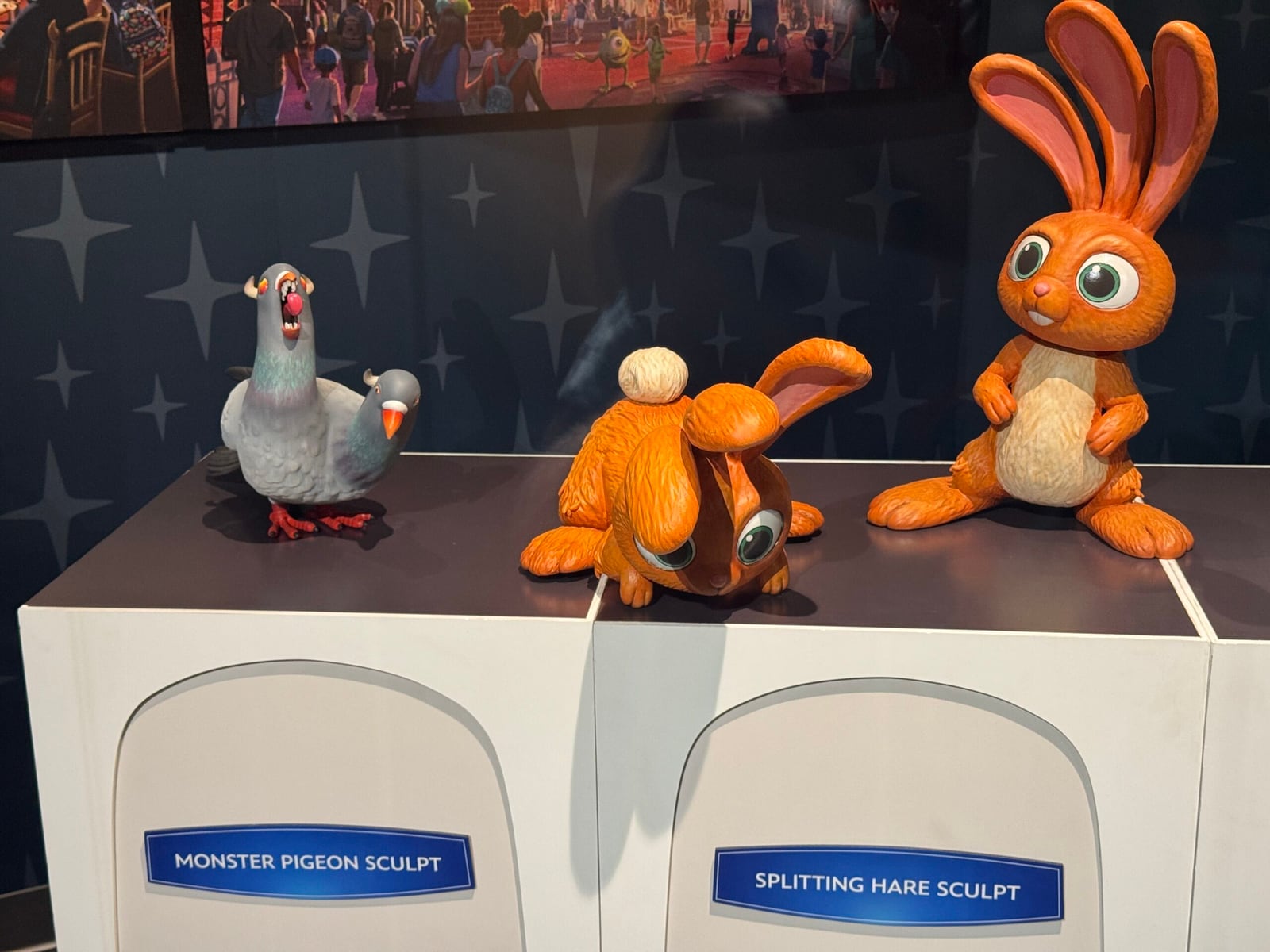 Three sculptures on white pedestals against a starry blue wall: a grey two-headed pigeon with one head screaming, an orange hare curled up with its head split in two, and a second orange hare standing upright with big eyes, above blue signs reading Monster Pigeon Sculpt and Splitting Hare Sculpt