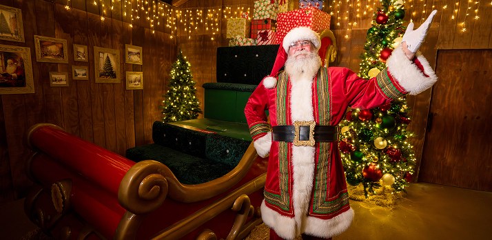 Santa Claus in a red suit with green and gold trim and a gold-buckled black belt, waving a white-gloved hand beside a red sleigh in a wood-paneled cabin set with lit Christmas trees, a stack of wrapped gifts and framed winter pictures on the wall