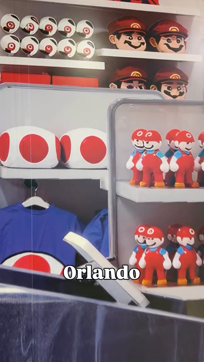 Another section of the construction wall graphic: shelves of red and white spotted plush, rows of small red-capped figures in red outfits with blue straps, rows of mustached, Mario-style faces in red caps, and a blue shirt with a red and white spot design, with the word Orlando captioned over the frame