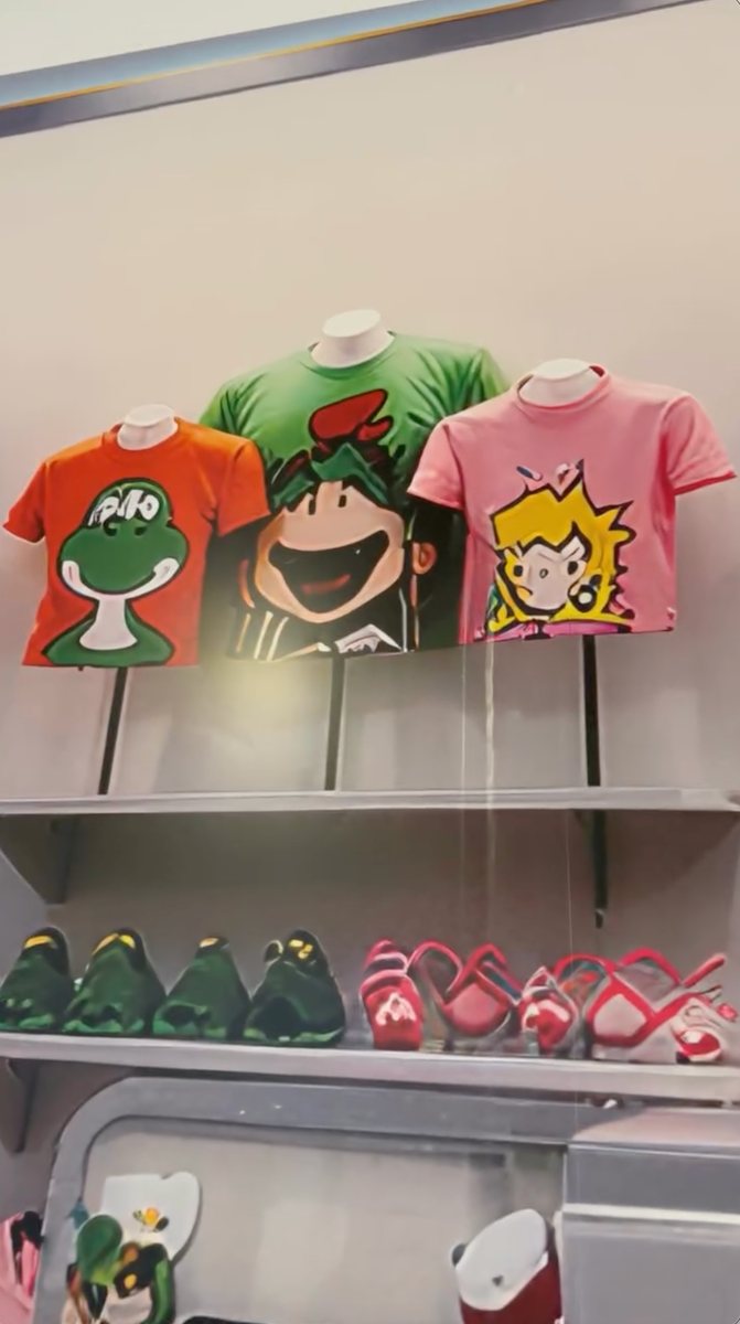 A construction wall graphic showing three T-shirts on display busts: an orange shirt with a distorted Yoshi and unreadable lettering, a green shirt with a warped Mario-style face, and a pink shirt with a lopsided Princess Peach, above a shelf of green and red shoe-like shapes