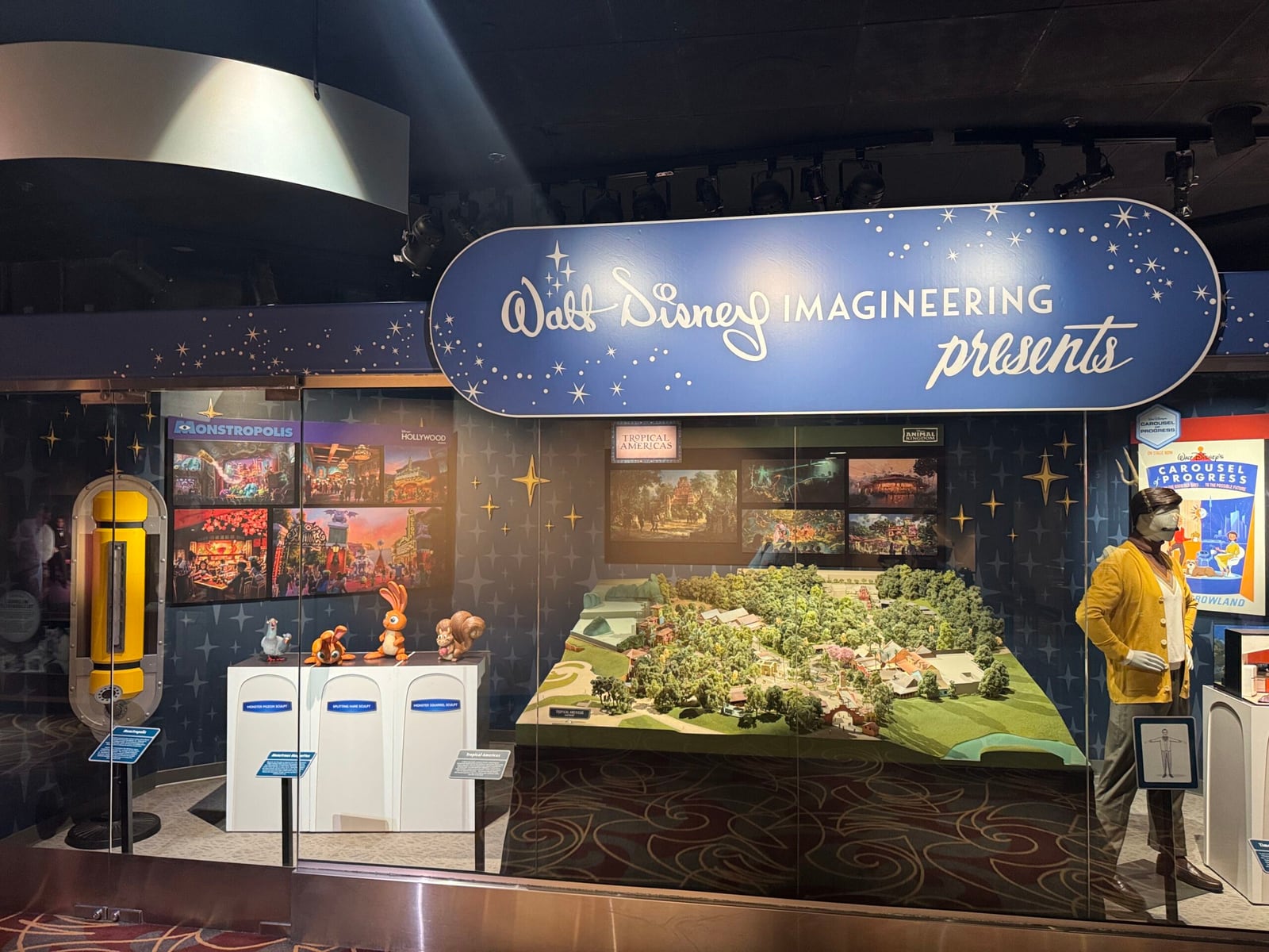 A wide view of a darkened exhibit room under a Walt Disney Imagineering Presents sign: Monstropolis concept art and a yellow canister at left, creature sculptures on white pedestals, a large landscaped scale model in the center, and a mannequin in a yellow cardigan at right