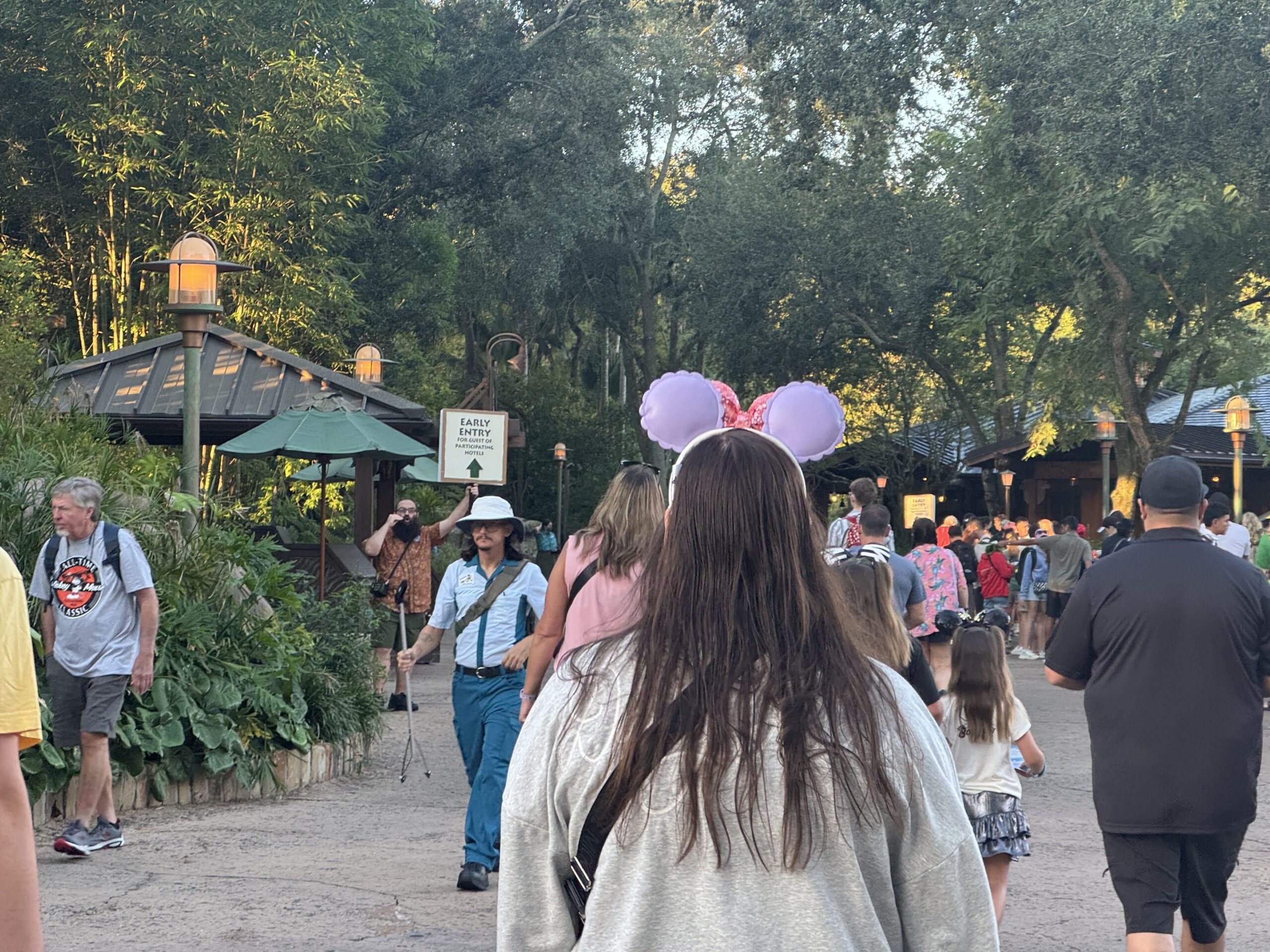 Guests walking along a tree-lined path at Disney's Animal Kingdom in early morning light, past a sign held aloft reading Early Entry for guest of participating hotels with a green arrow pointing ahead, while a crowd gathers under a second Early Entry sign at right