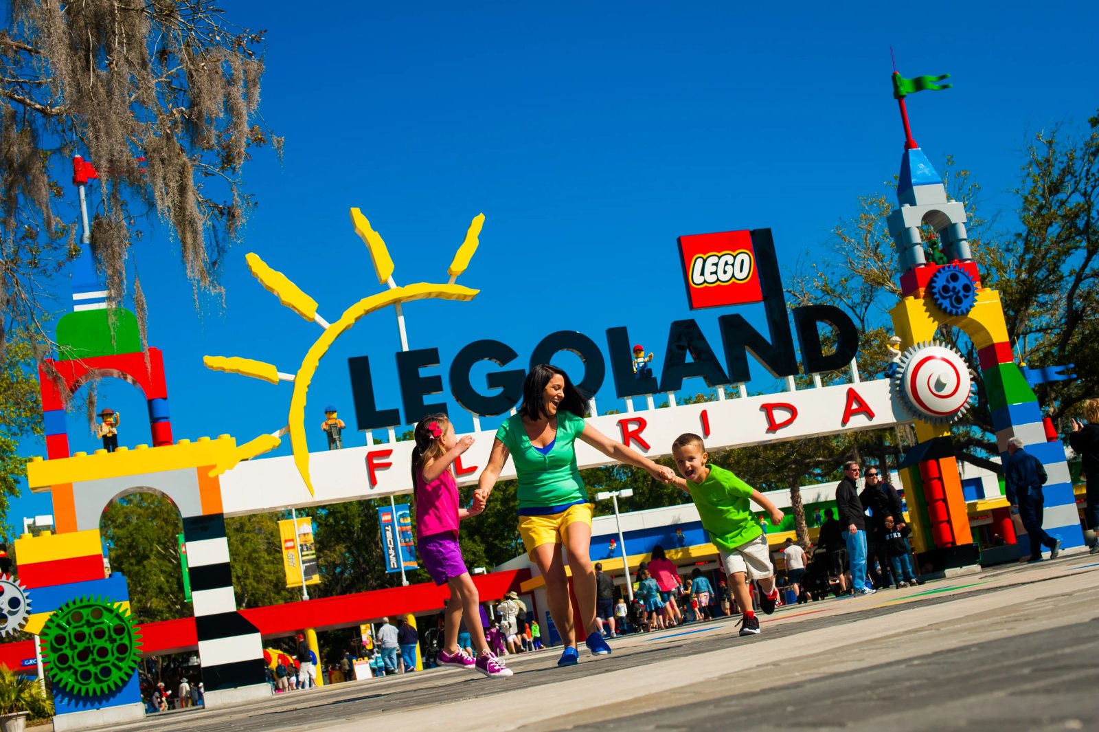 A smiling woman runs hand in hand with a girl and a boy past the colorful LEGO brick entrance arch reading LEGOLAND FLORIDA, with a yellow LEGO sun and LEGO logo on top