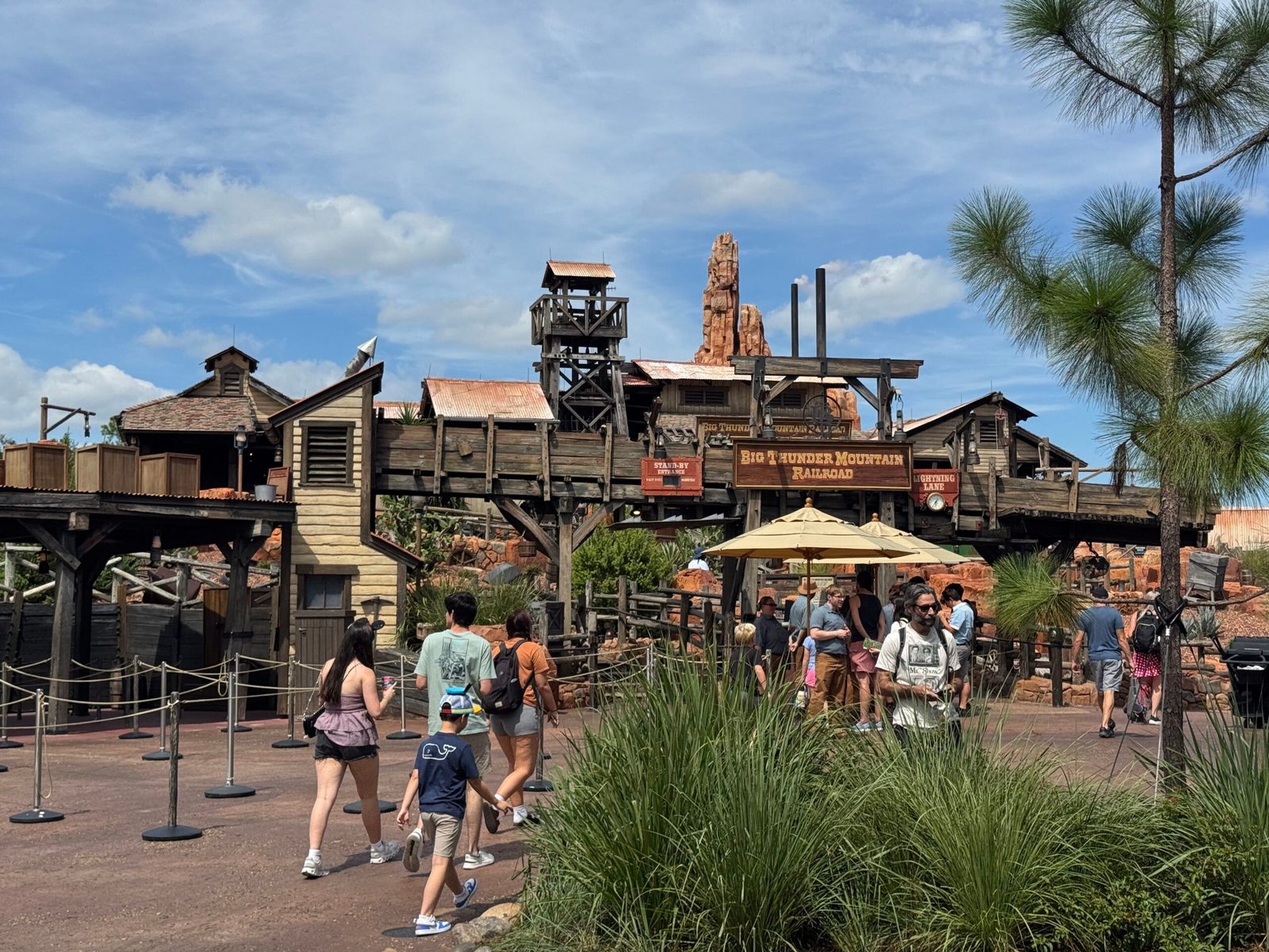 The Big Thunder Mountain Railroad entrance in Frontierland, with the ride's wooden sign, a standby entrance sign and a Lightning Lane sign, and a group of people, including cast members, gathered by two umbrellas at the queue entrance while guests walk past