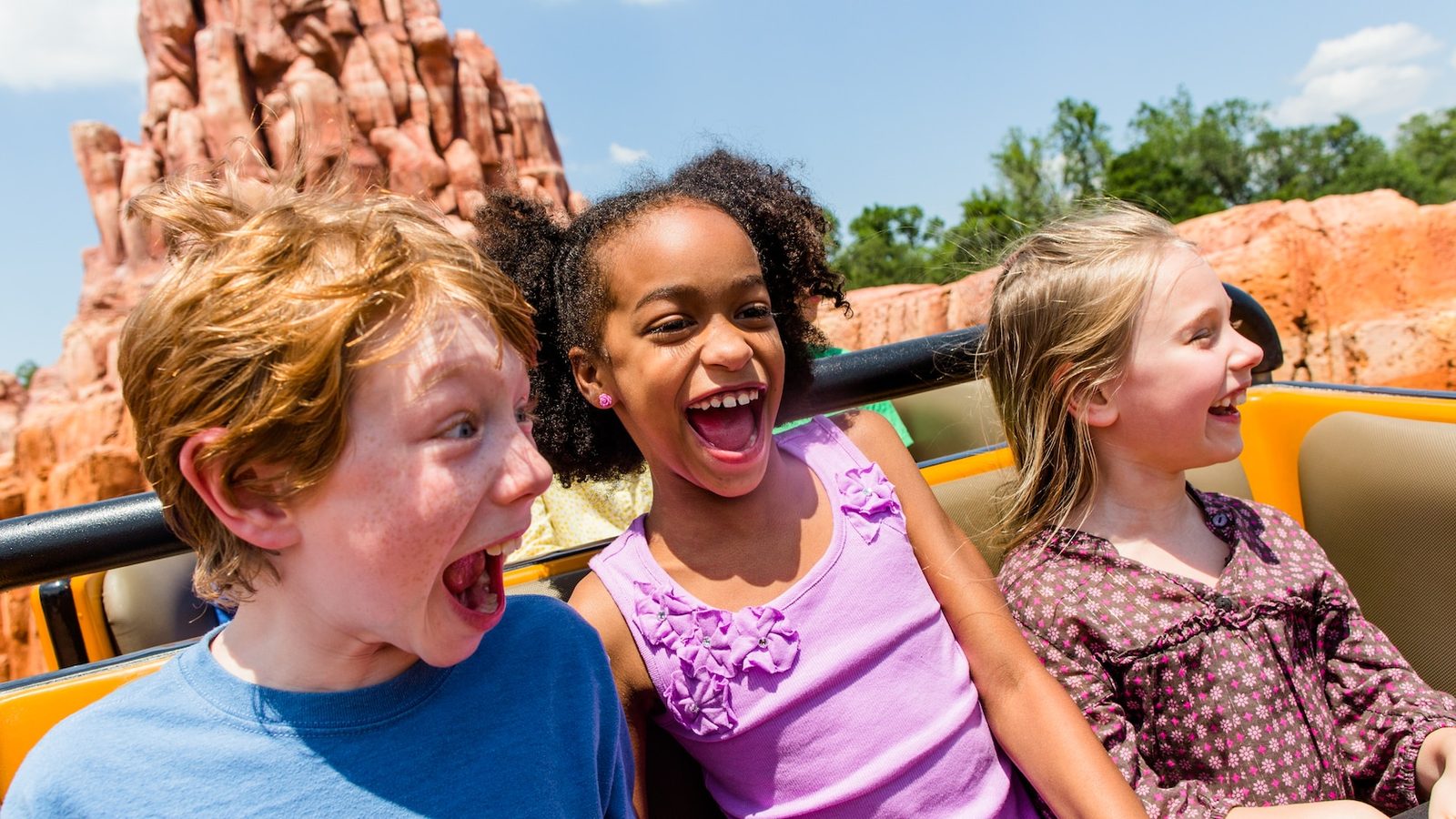 Three children laughing and shouting in an open Big Thunder Mountain Railroad train car, with the ride's red rock spires behind them under a blue sky
