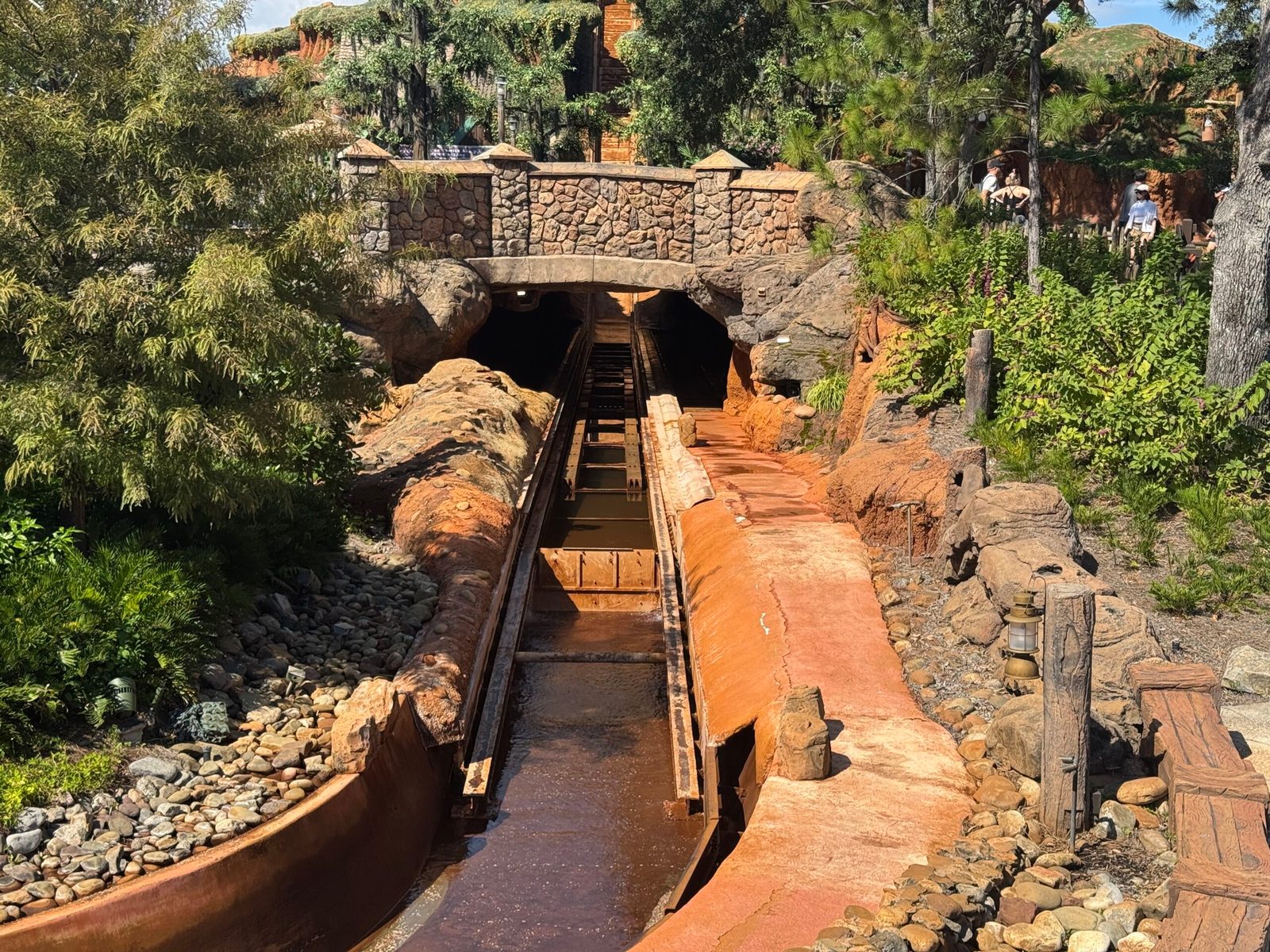 A stretch of the Tiana's Bayou Adventure flume running under a stone bridge, with the channel holding only a shallow layer of brown water so the track and guide rails at the bottom are exposed