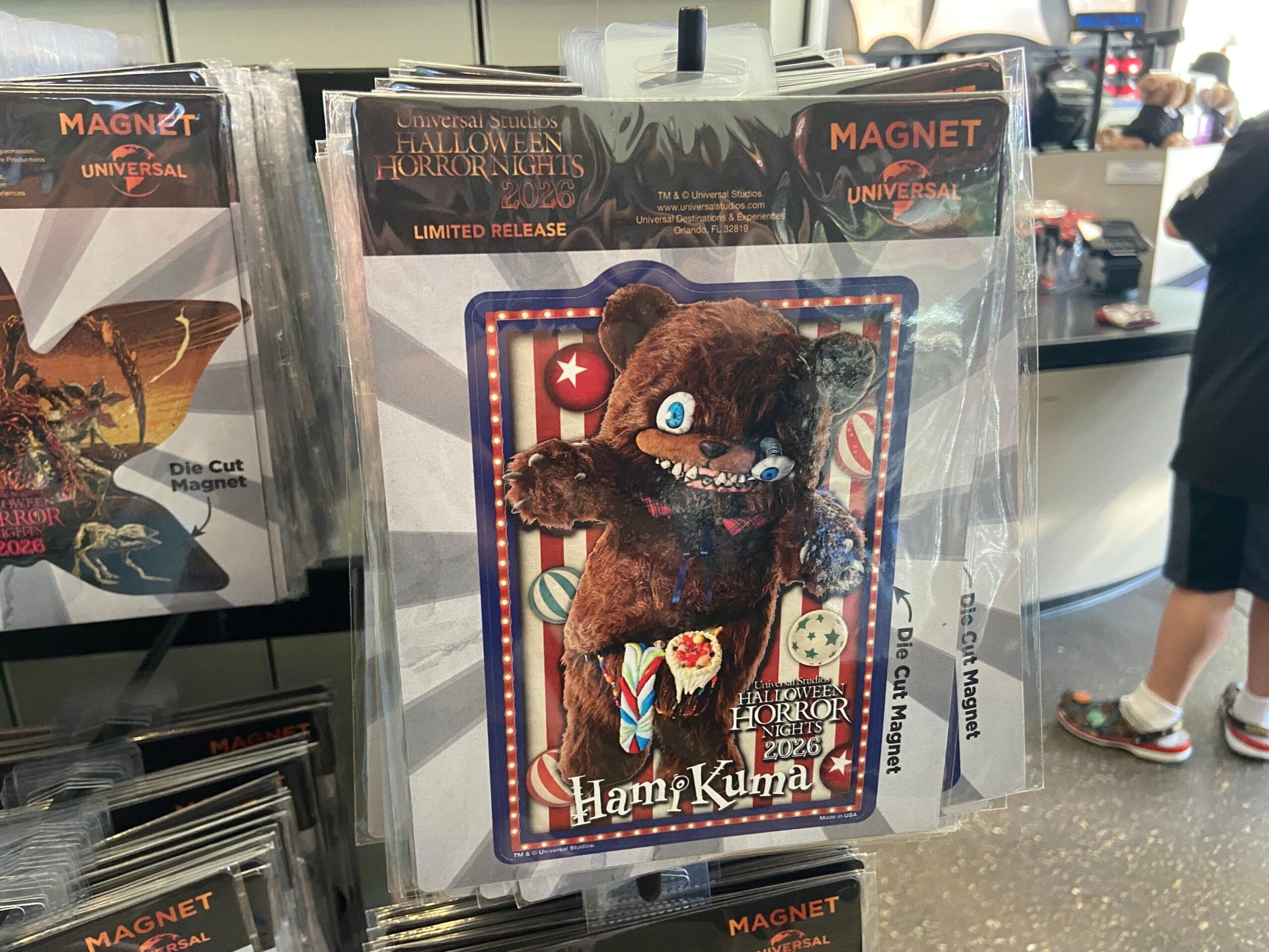 A packaged die-cut magnet showing a brown zombie teddy bear against a red and white striped carnival background, with the name HamiKuma and a Halloween Horror Nights 2026 logo