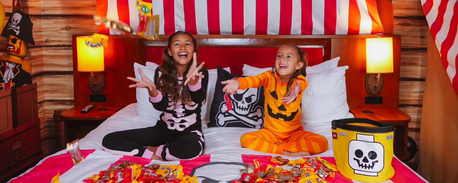 Two laughing girls in skeleton and jack-o'-lantern pajamas toss candy on a bed in a pirate-themed hotel room, with a yellow LEGO skull-head treat bucket beside them