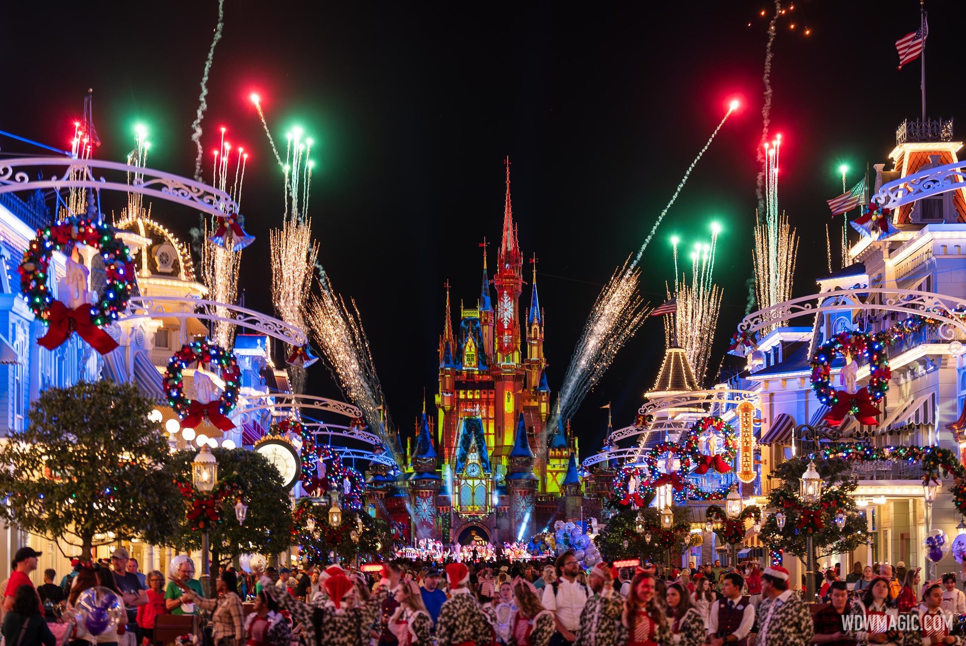 Red and green fireworks rise along a wreath-hung Main Street, U.S.A. toward Cinderella Castle, lit with red and gold holiday projections, as crowds fill the street