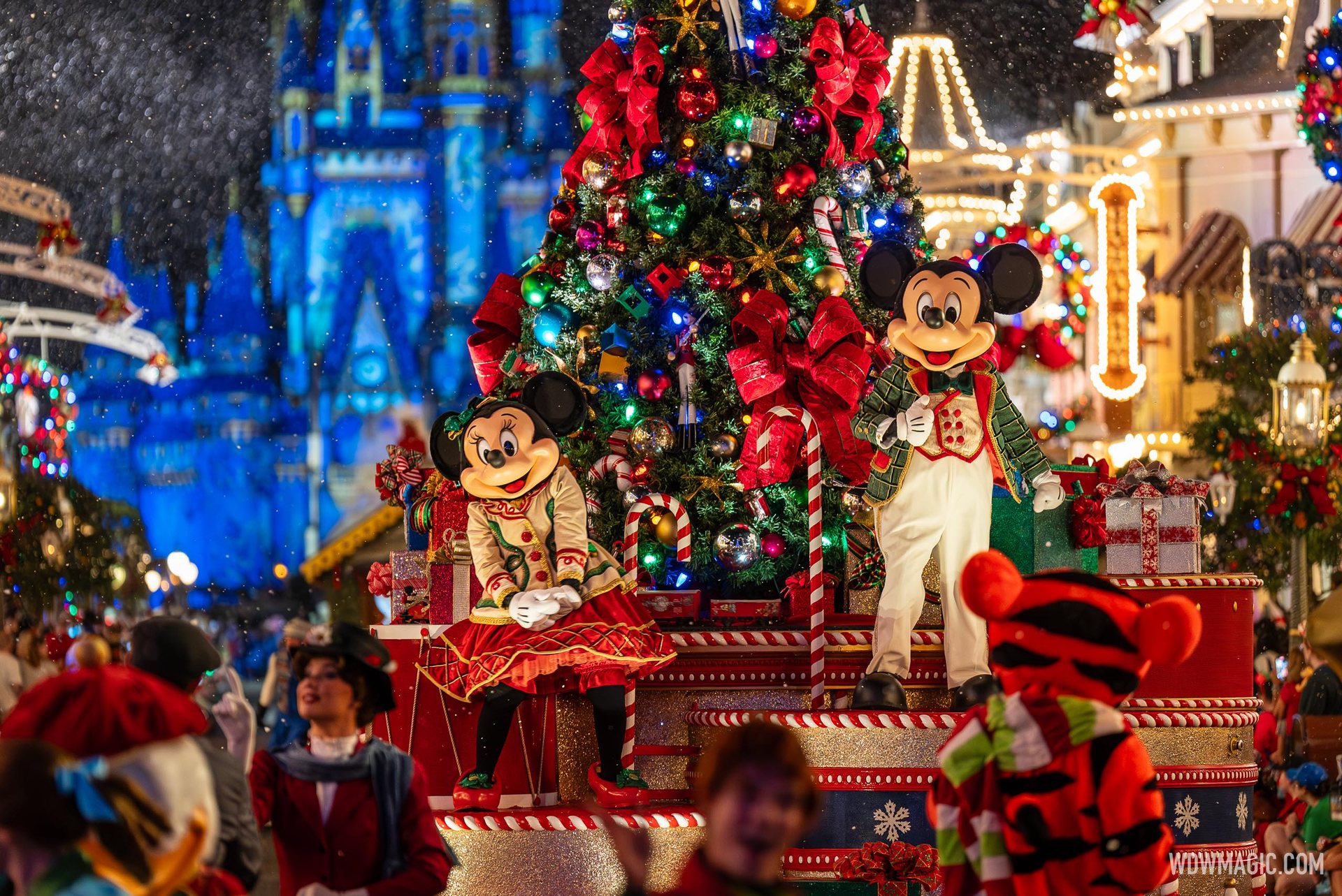 Mickey and Minnie Mouse in festive outfits ride a parade float beside a decorated Christmas tree, with Cinderella Castle lit blue behind them