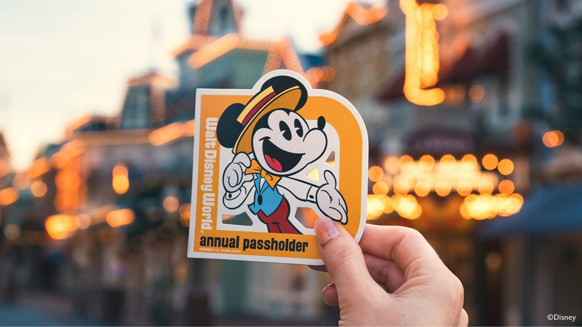 A hand holds up a die-cut golden-yellow Walt Disney World annual passholder magnet showing Mickey Mouse in a straw boater hat, yellow bow tie, blue vest and red trousers, tipping his hat, in front of a blurred Main Street, U.S.A. lit with warm bulbs