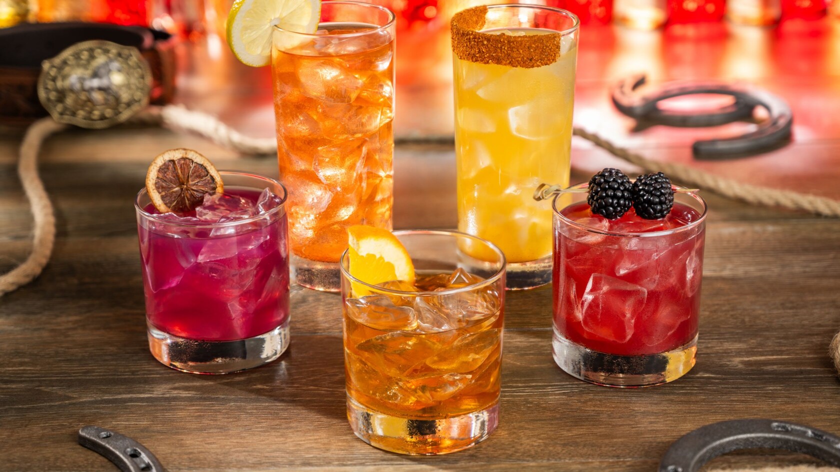 Five cocktails on a wooden table with rope, horseshoes and a belt buckle: a purple drink with a dried citrus wheel, a tall amber drink with a lemon wheel, a tall yellow drink with a spiced rim, an amber drink on ice with an orange slice, and a red drink topped with blackberries