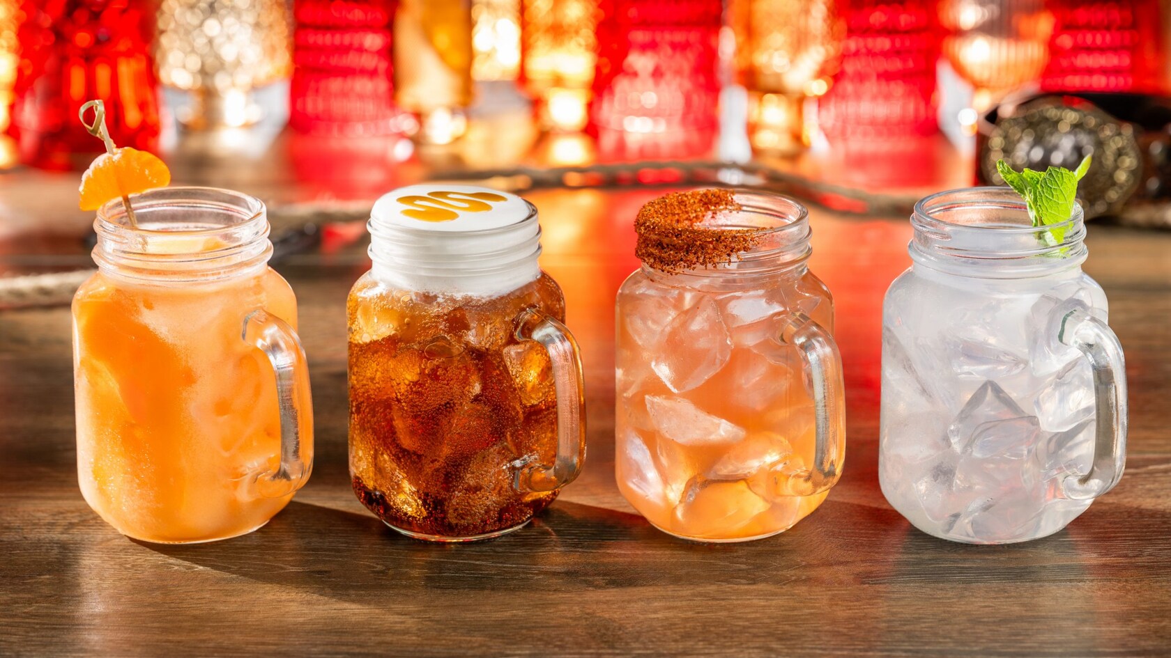 Four drinks in glass mason-jar mugs on a wooden bar: an orange drink with an orange garnish, a root beer topped with cream and a caramel swirl, a pink drink with a spiced rim, and a clear drink with mint