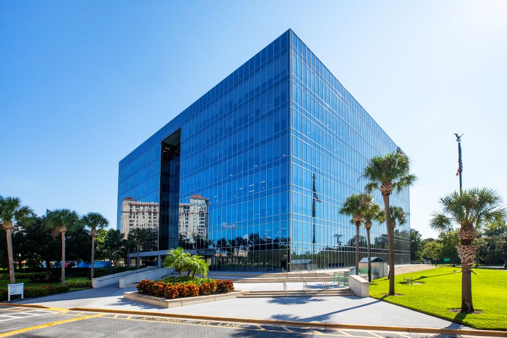 An eight-story blue glass cube office tower under a clear sky, with palm trees, a green lawn and a flagpole in front, and neighboring buildings reflected in its glass
