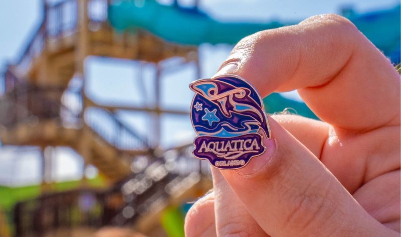 A hand holds up a purple and blue enamel Aquatica Orlando pin showing a Commerson's dolphin, in front of a blurred water slide tower