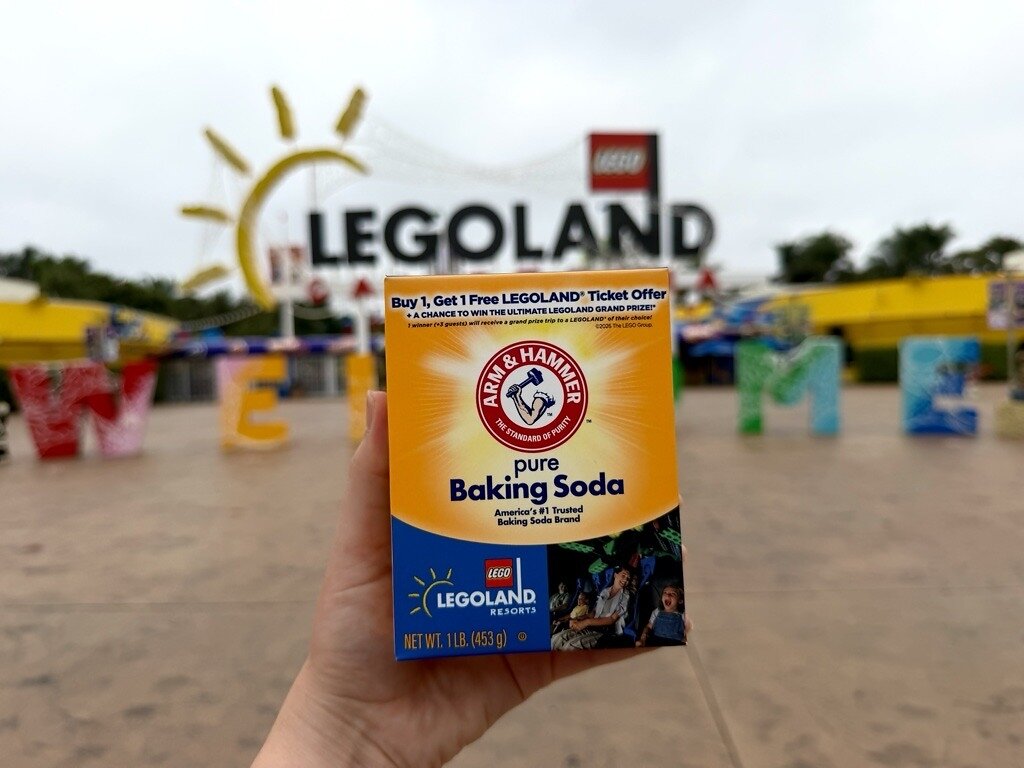 A hand holds an orange ARM & HAMMER Baking Soda box with LEGOLAND Resorts artwork and a Buy 1, Get 1 Free LEGOLAND Ticket Offer banner, in front of a LEGOLAND entrance sign