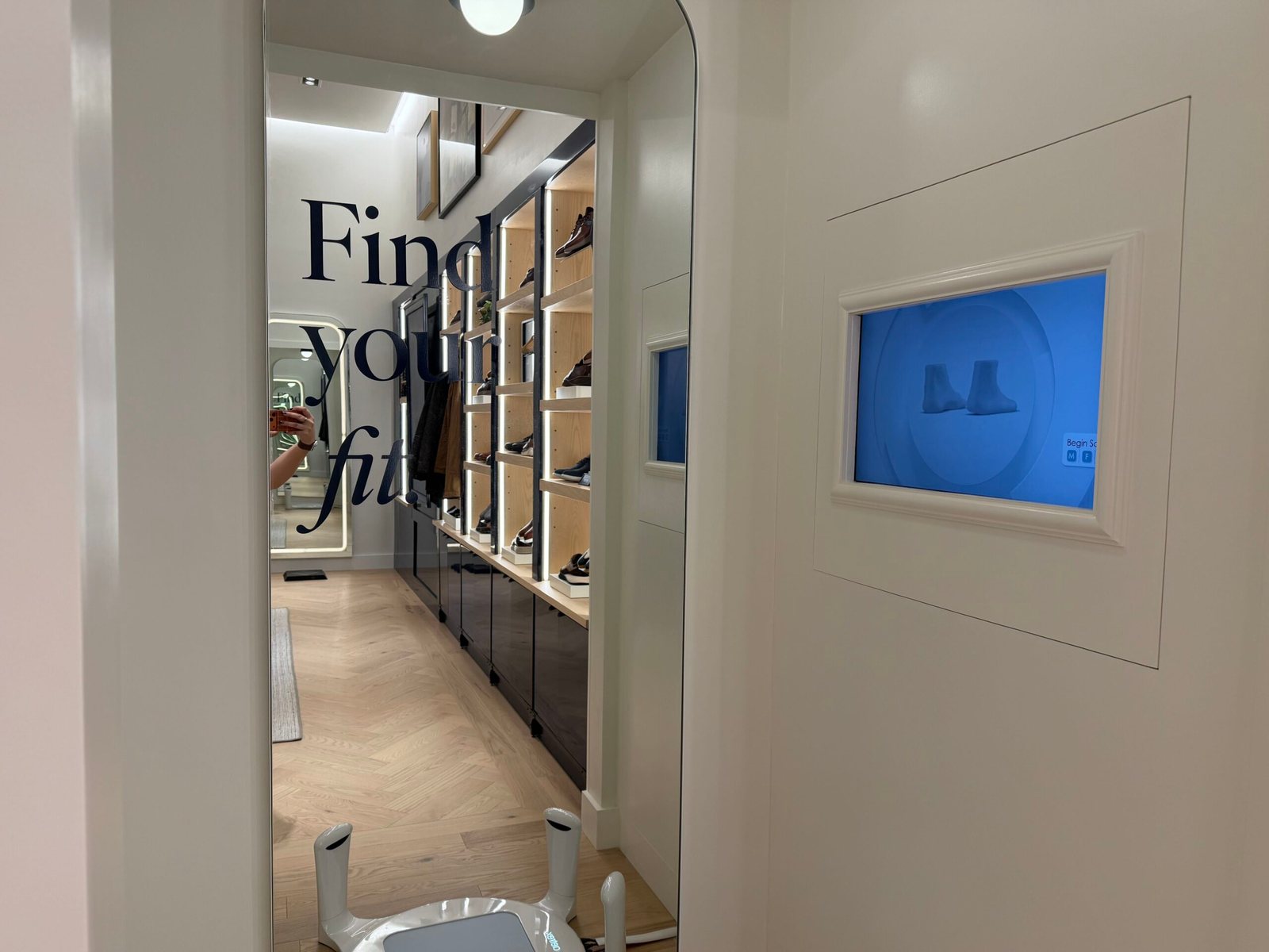 A mirror with Find your fit lettering beside a blue screen set into the wall showing a pair of 3D-rendered feet, with the white base of the foot scanner at the bottom of the frame