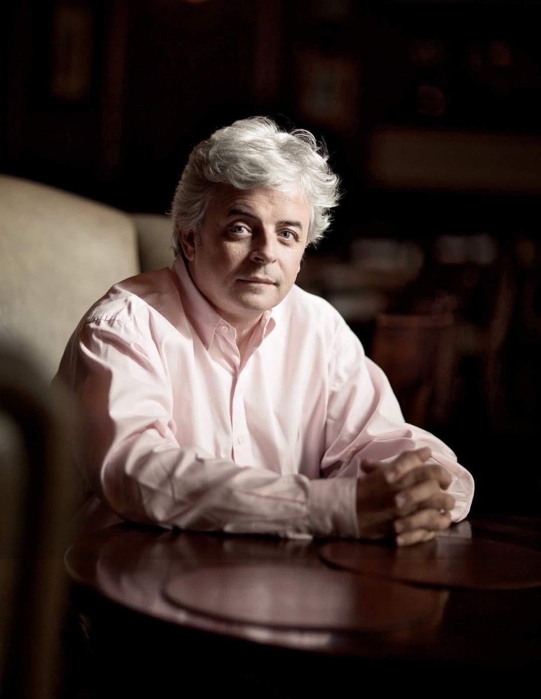 Paul Nolan in a pale pink shirt, seated at a dark wooden table with his hands clasped, in a dimly lit room