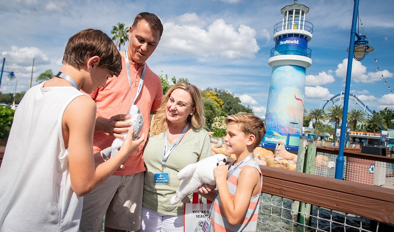 A family wearing Pass Member lanyards holds plush seal toys on a boardwalk, with the SeaWorld lighthouse and a coaster behind them
