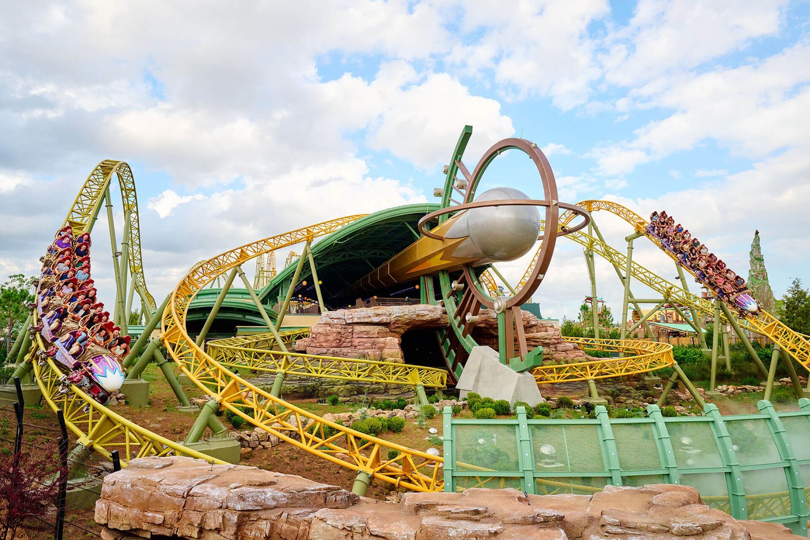 Two Stardust Racers trains full of riders on yellow and green coaster track in Celestial Park, with a silver comet sculpture circled by a large ring at the center