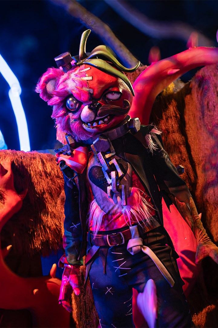 A costumed bear character with pink fur, a reddish-orange stitched face, a metal bolt and curled green cap on its head, mismatched eyes and a stitched jacket stands in front of a straw wall under blue neon