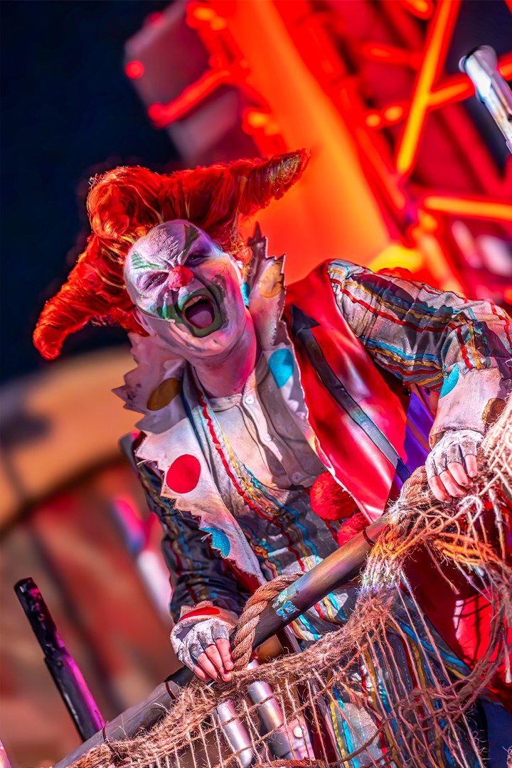 Jack the Clown, with green and white face paint, swept red hair and a polka-dot costume, screams while gripping a rope-wrapped podium railing in front of red neon
