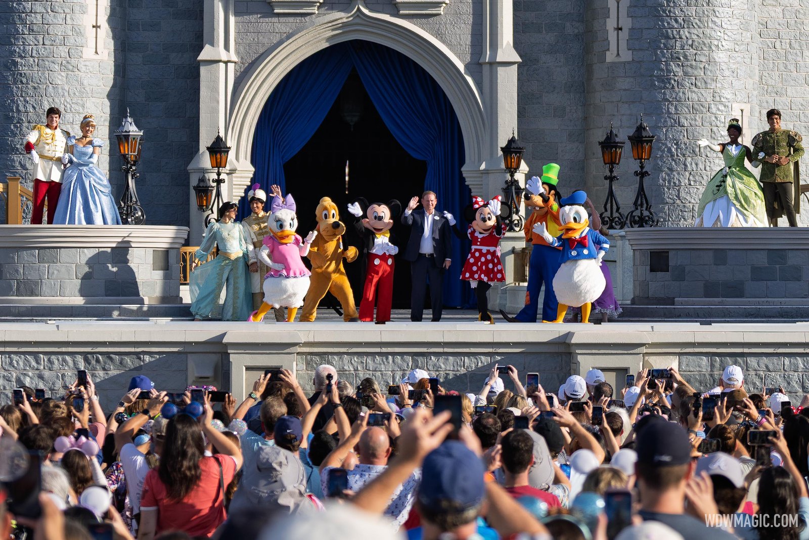 Joe Schott stands at center of the Cinderella Castle stage between Mickey and Minnie, with Donald, Daisy, Goofy, Pluto and Disney princes and princesses on the stage and upper balconies in front of blue curtains, and a crowd holding up phones below