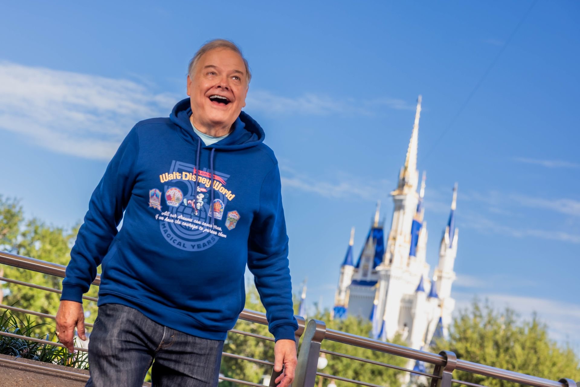 Opening day cast member Frank, smiling in a blue Walt Disney World 55 Magical Years hoodie, walks beside a railing with Cinderella Castle behind him