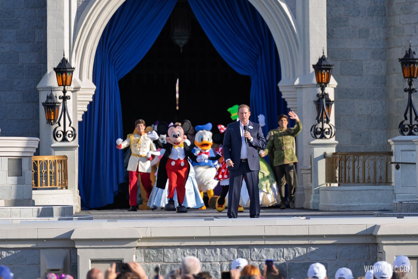 Joe Schott in a navy suit speaks into a microphone on the Cinderella Castle stage as Mickey, Donald, Prince Charming and other characters walk out through blue curtains behind him